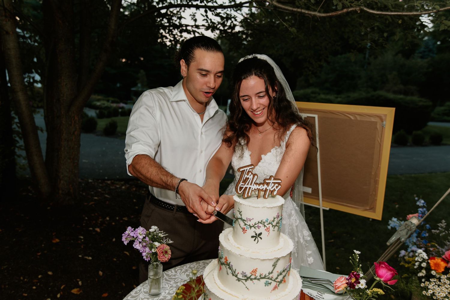 Bride and groom cutting a floral white wedding cake outdoors at dusk.