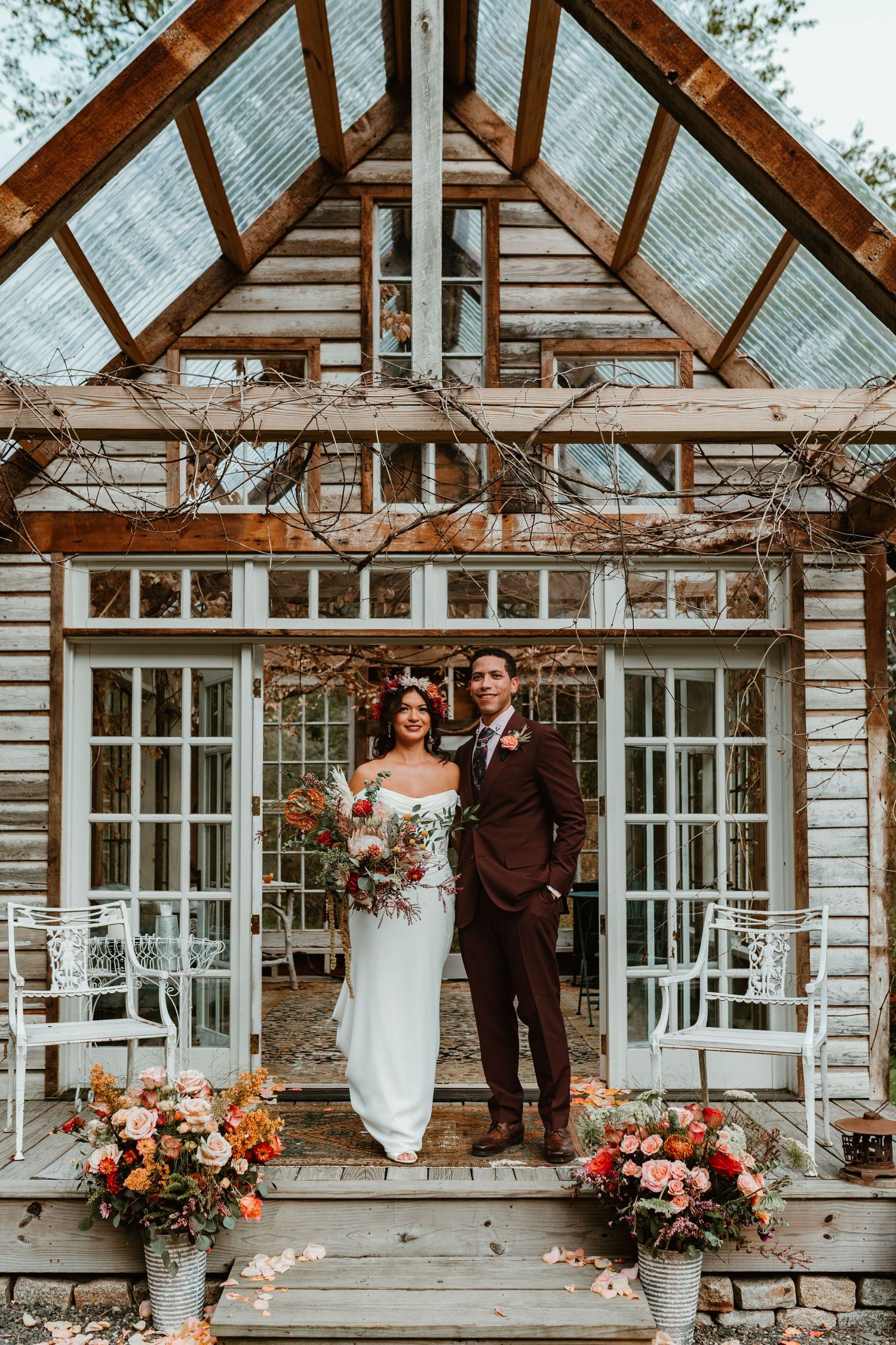 A couple stands on a rustic greenhouse porch decorated with floral arrangements for their wedding day.