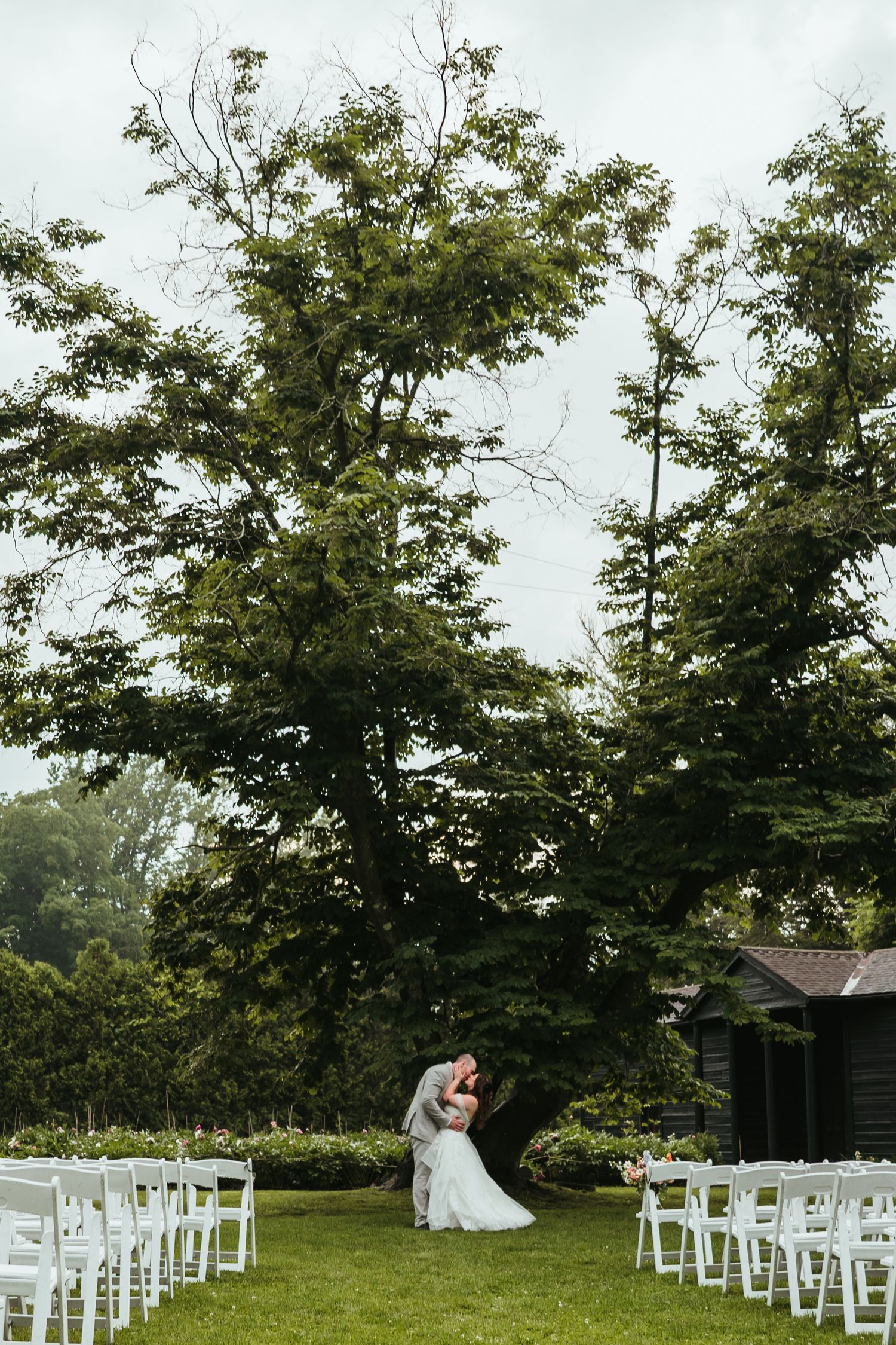 White chairs arranged for an outdoor wedding ceremony under tall trees in a serene garden setting.