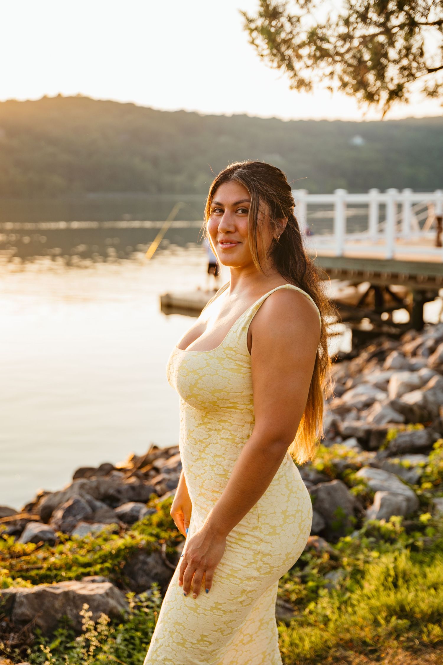 Woman in yellow dress posing by rocky lakeside at golden hour sunset with dock in background.
