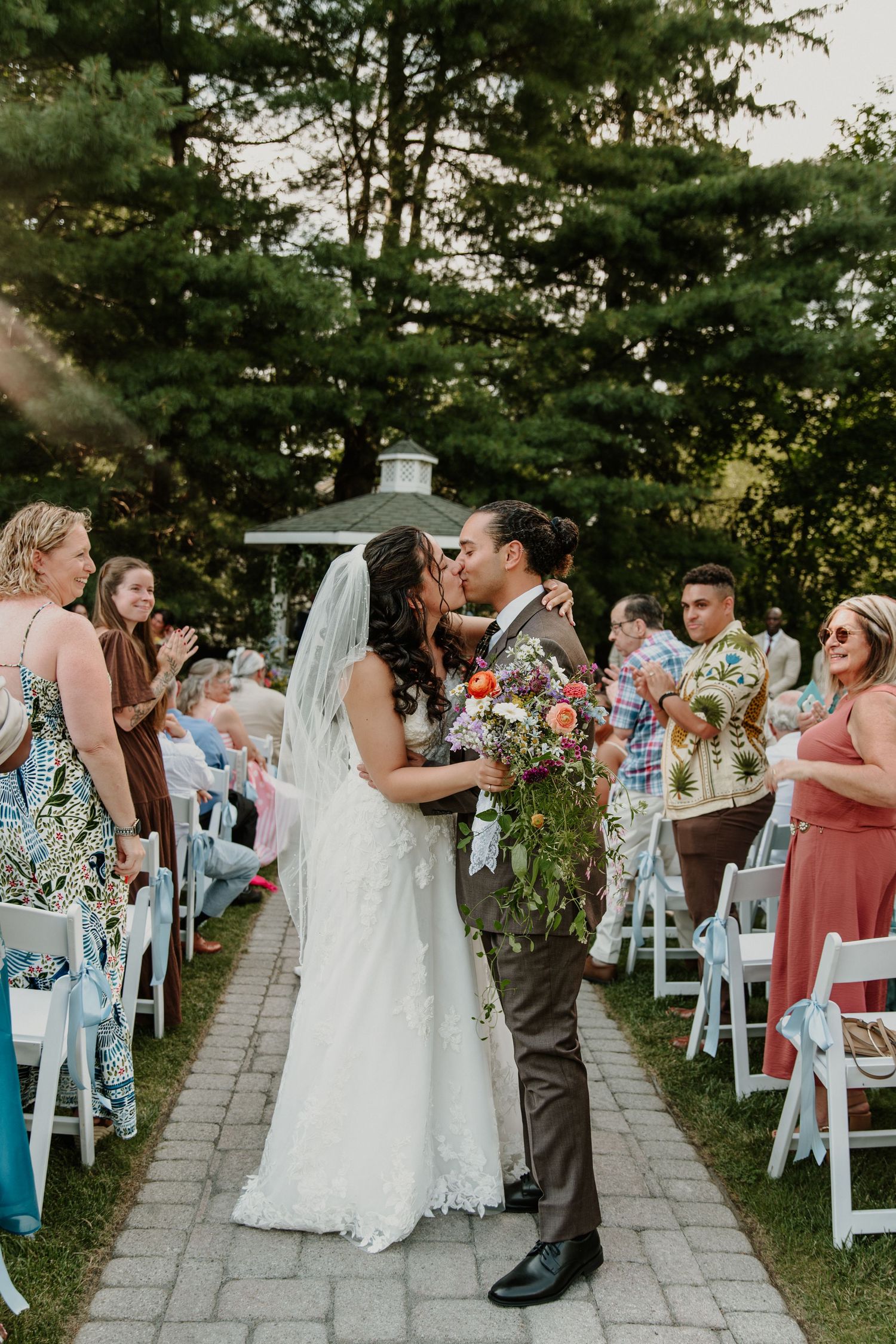 Bride and groom share first kiss as newlyweds while walking down outdoor aisle lined with guests in white chairs.