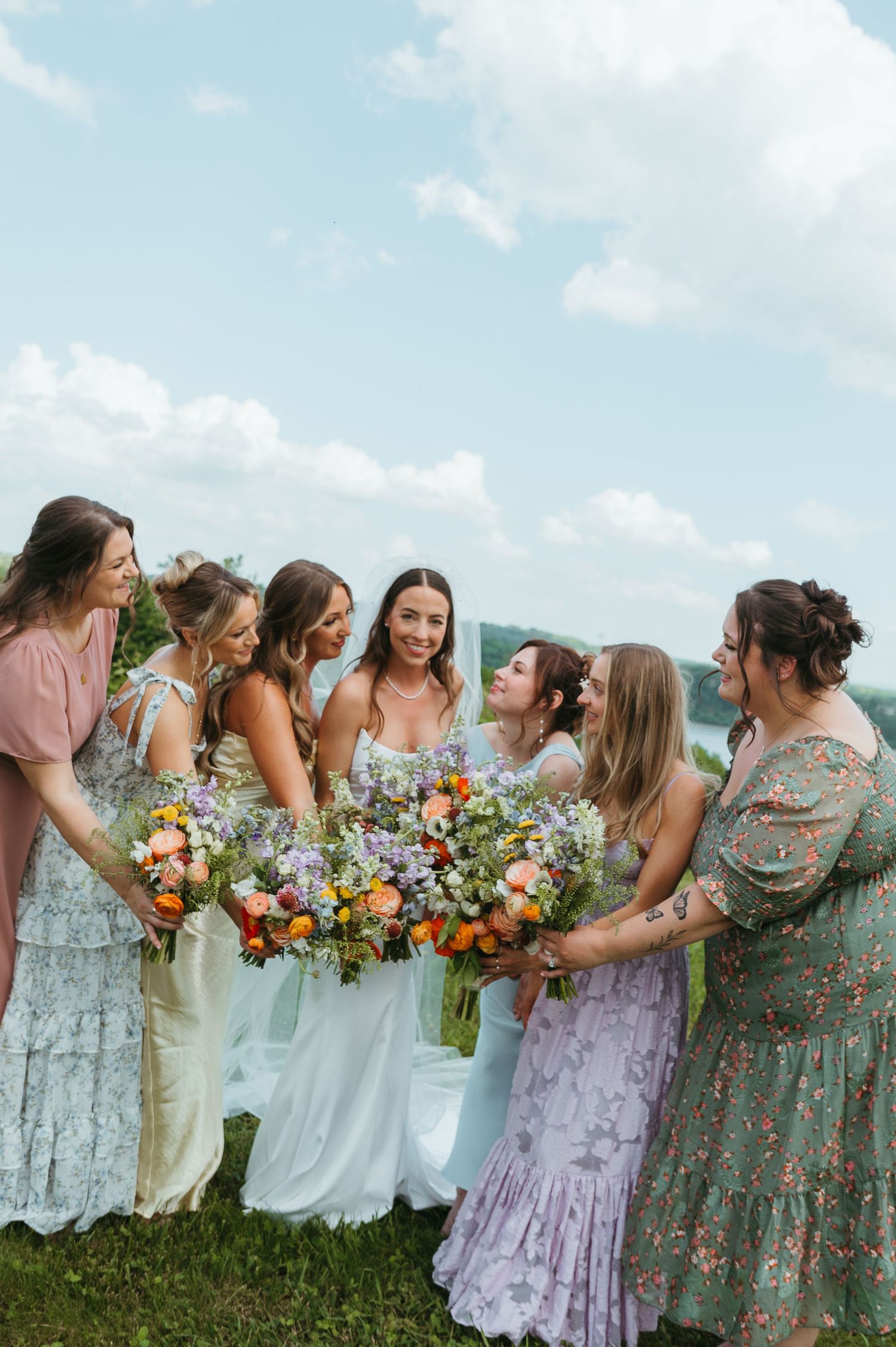Bridesmaids in floral pastel dresses hold colorful bouquets while posing against a blue sky.