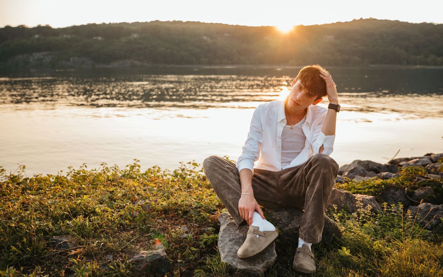 Person sitting on rocks by a sunlit lake at golden hour, wearing white shirt and casual pants outdoors.
