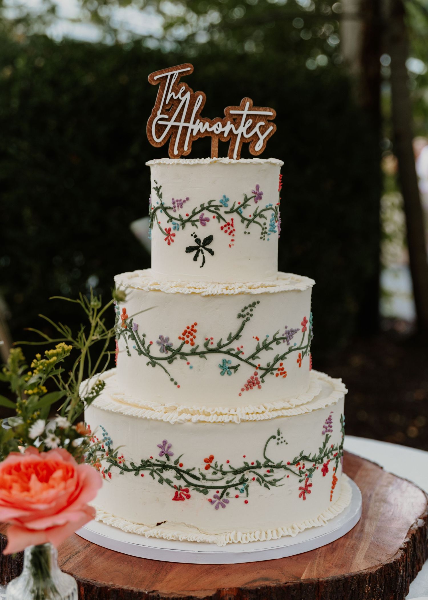 Three-tier white wedding cake with hand-painted floral vines and The Almontes wooden cake topper on rustic wood stand.