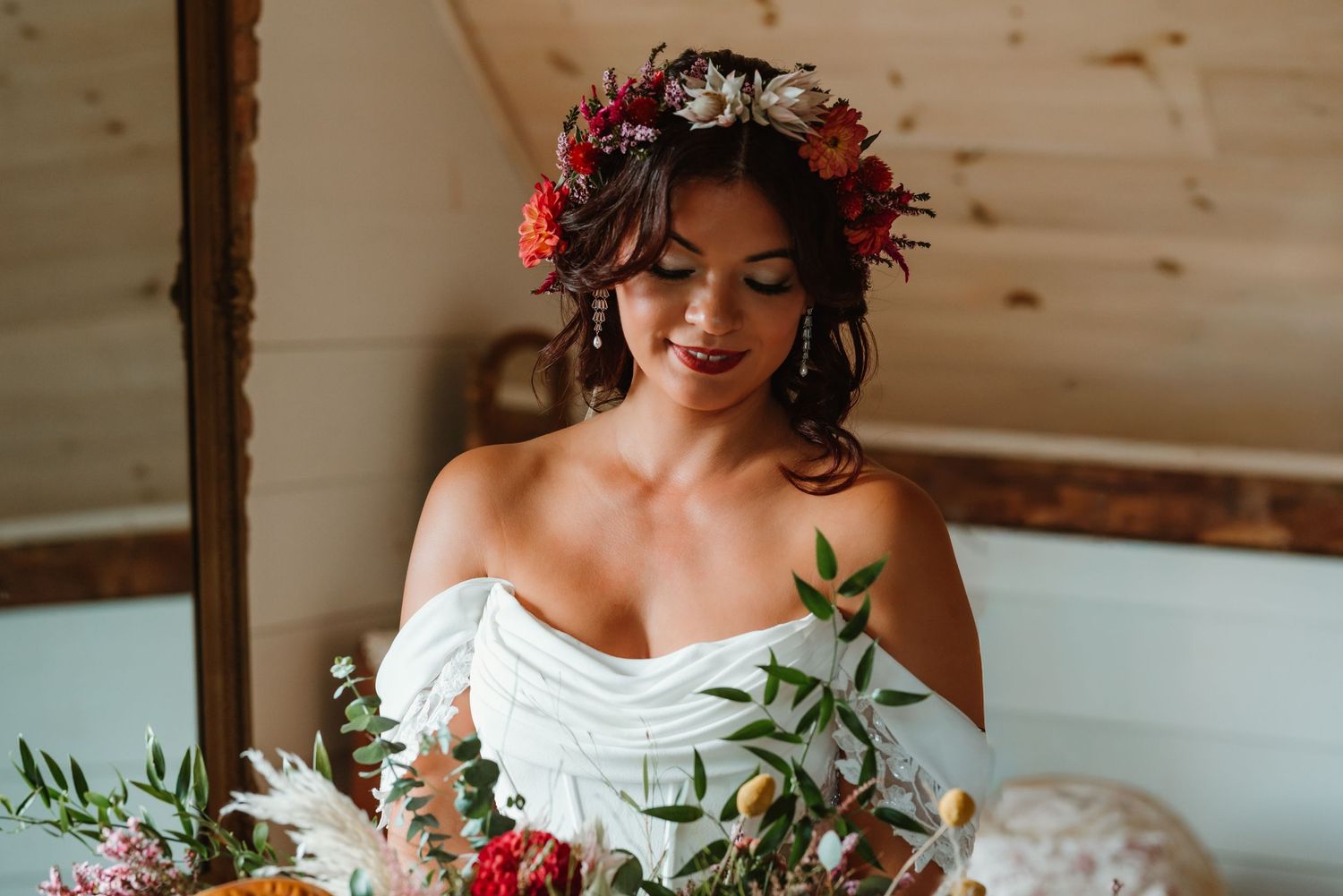 Bride wearing floral crown and off-shoulder dress poses with delicate bouquet in rustic indoor setting.