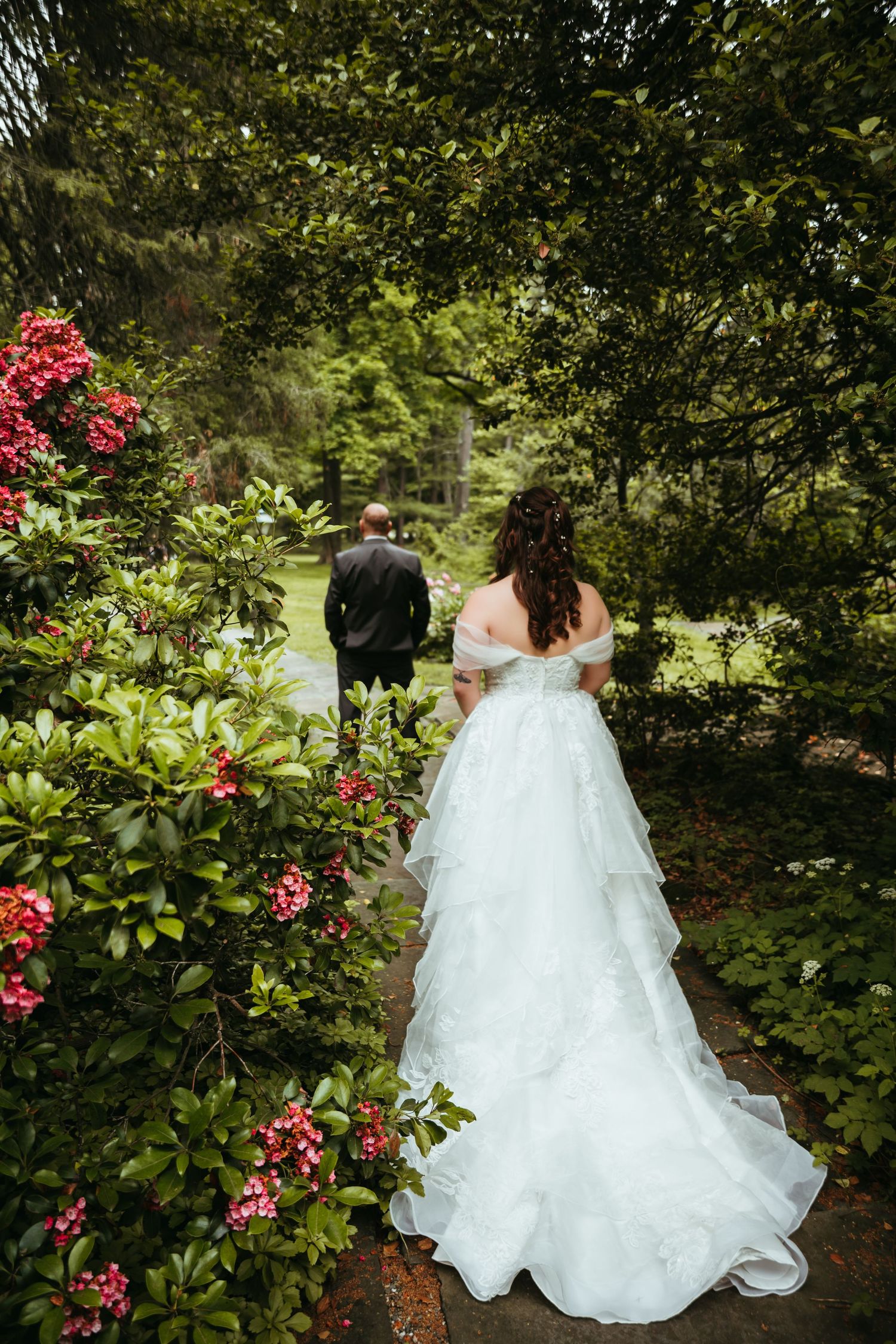 A couple in formal wedding attire walks through a lush garden path surrounded by blooming red roses and greenery.
