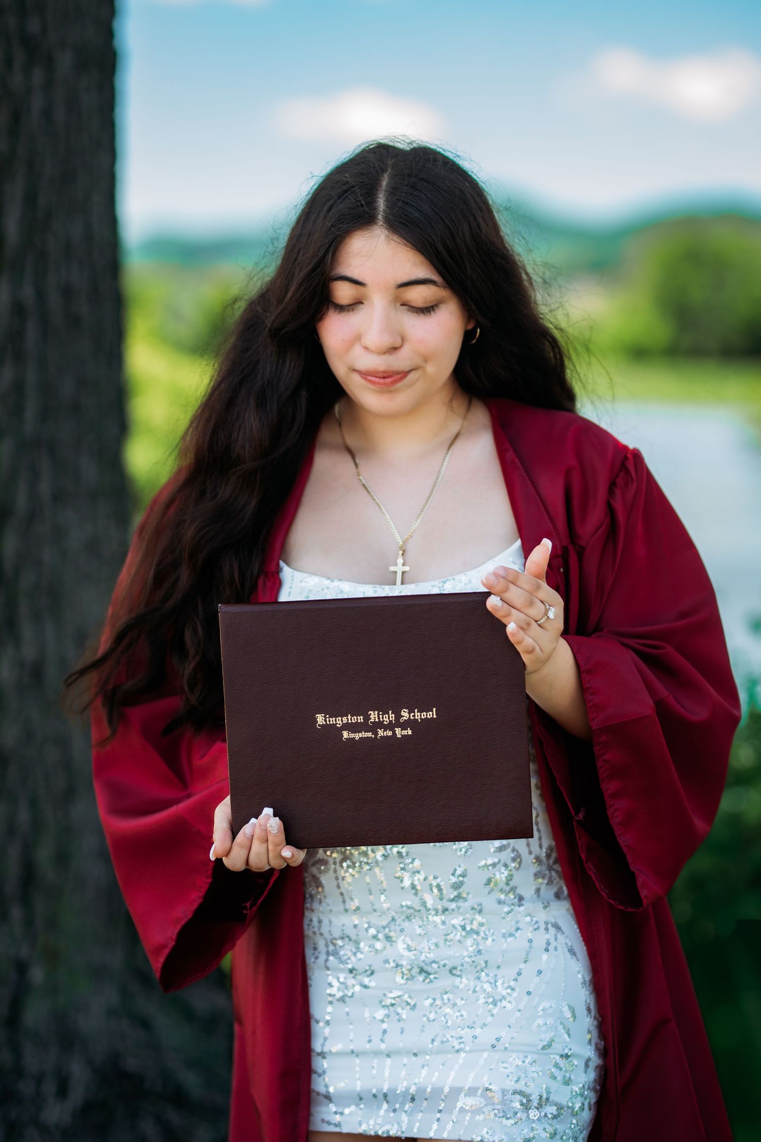 Young woman in red graduation gown holding Kingston High School diploma outdoors, smiling proudly at her achievement.