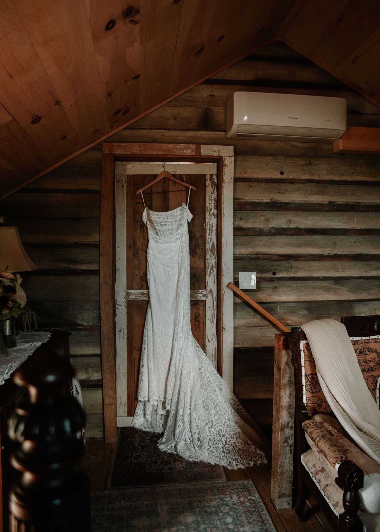 A white lace wedding dress hanging in a rustic log cabin with wooden walls and ceiling.