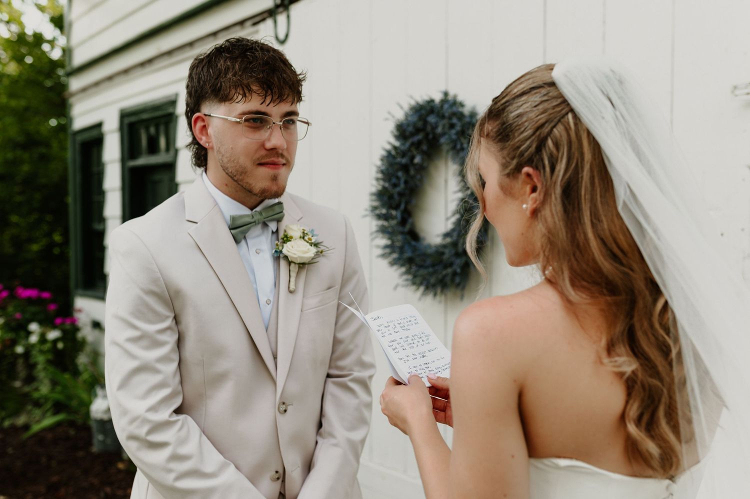 Groom in a beige suit and green bow tie listens as the bride in a veil reads her handwritten vows outdoors beside a white building.