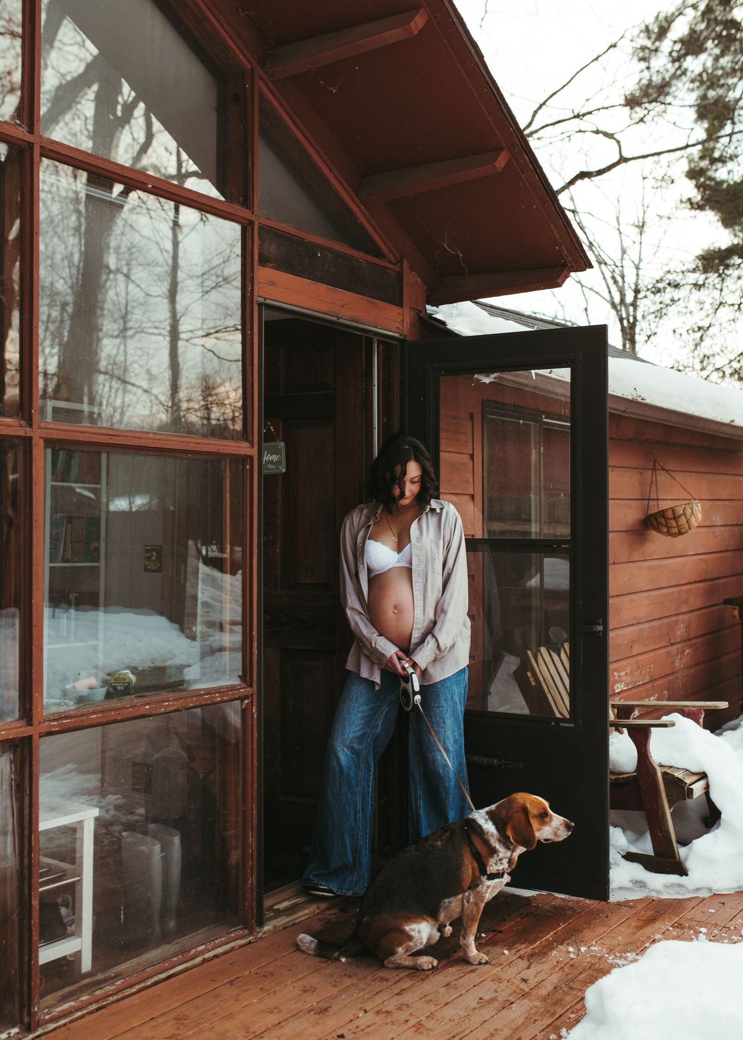 Pregnant woman in white bra and jeans standing in doorway of rustic wooden cabin with beagle dog in snow.