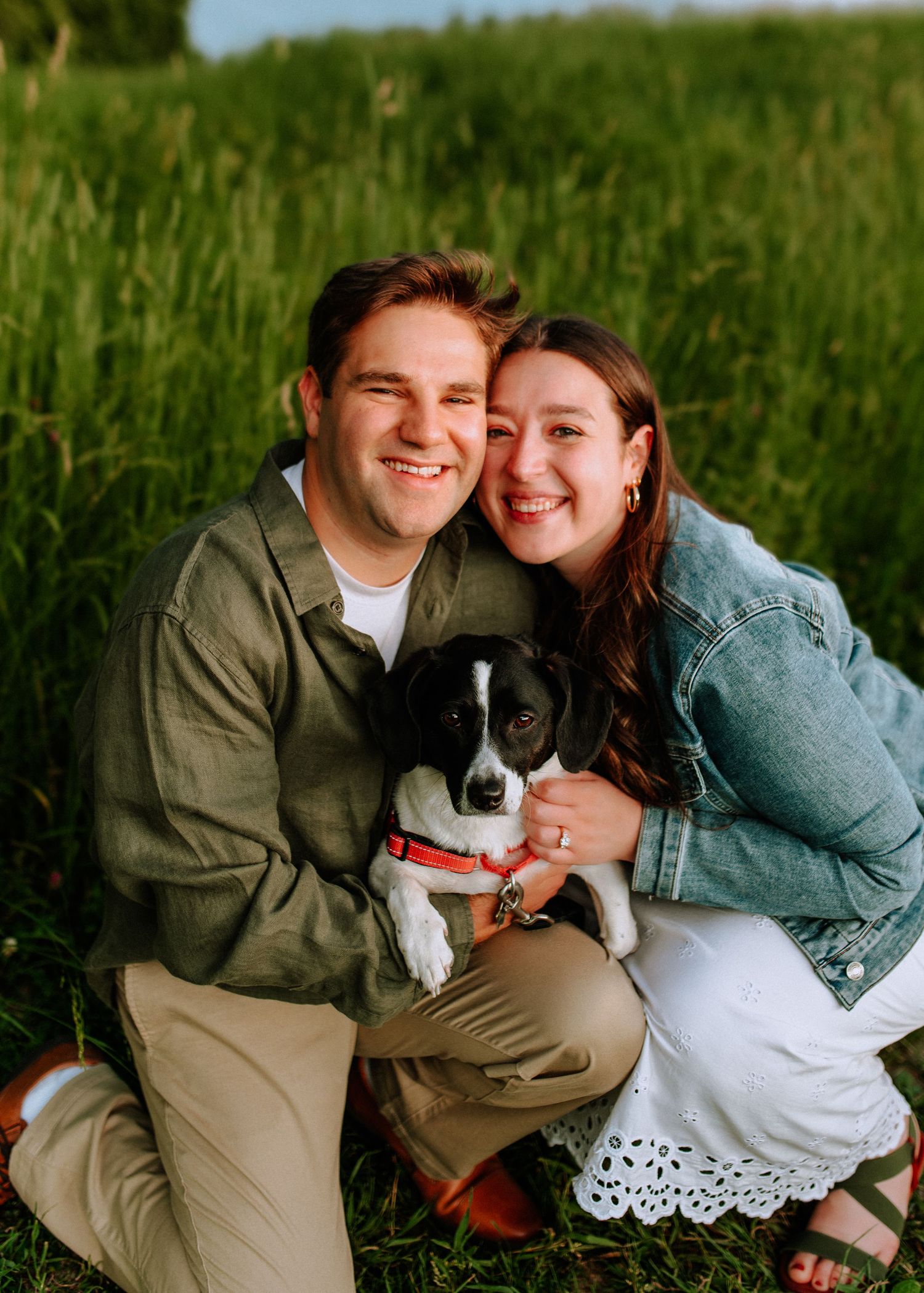Smiling couple poses outdoors in a green field holding a black and white puppy with a red collar.
