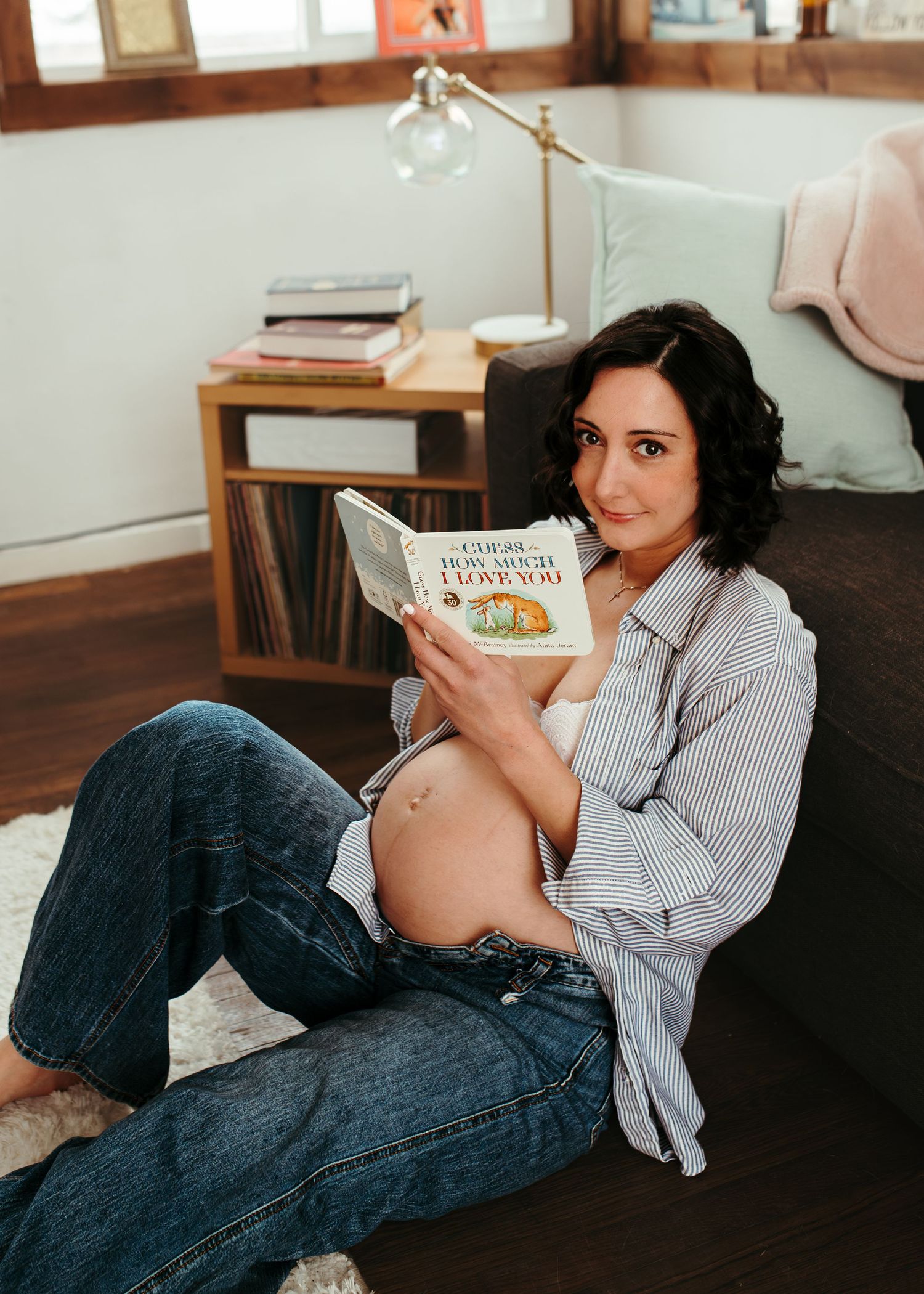 Pregnant woman sitting on floor reading 'Guess How Much I Love You' book, wearing jeans and striped shirt.