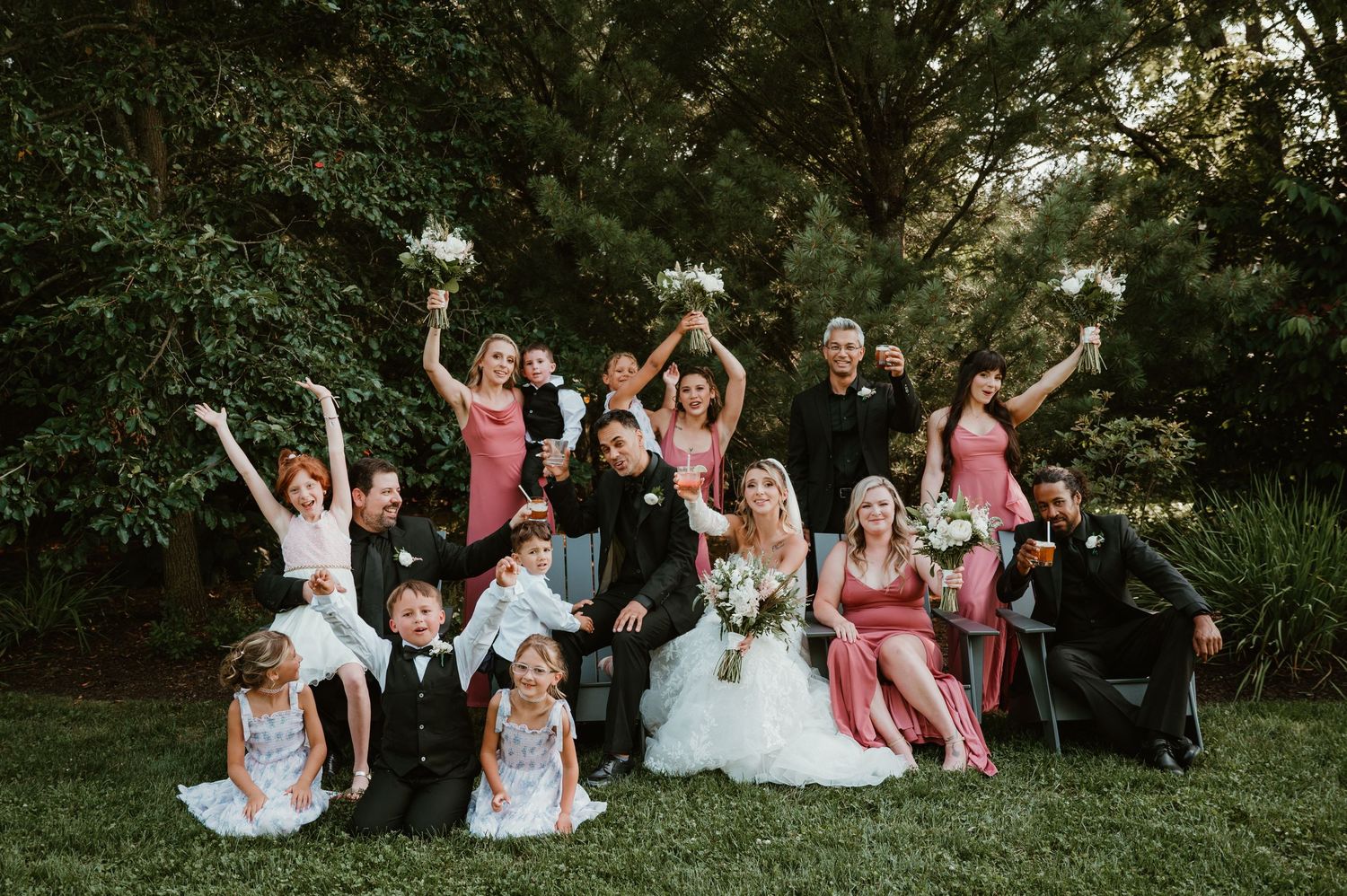 A wedding party sits in a grassy garden setting wearing pink and white attire while posing playfully for photos.