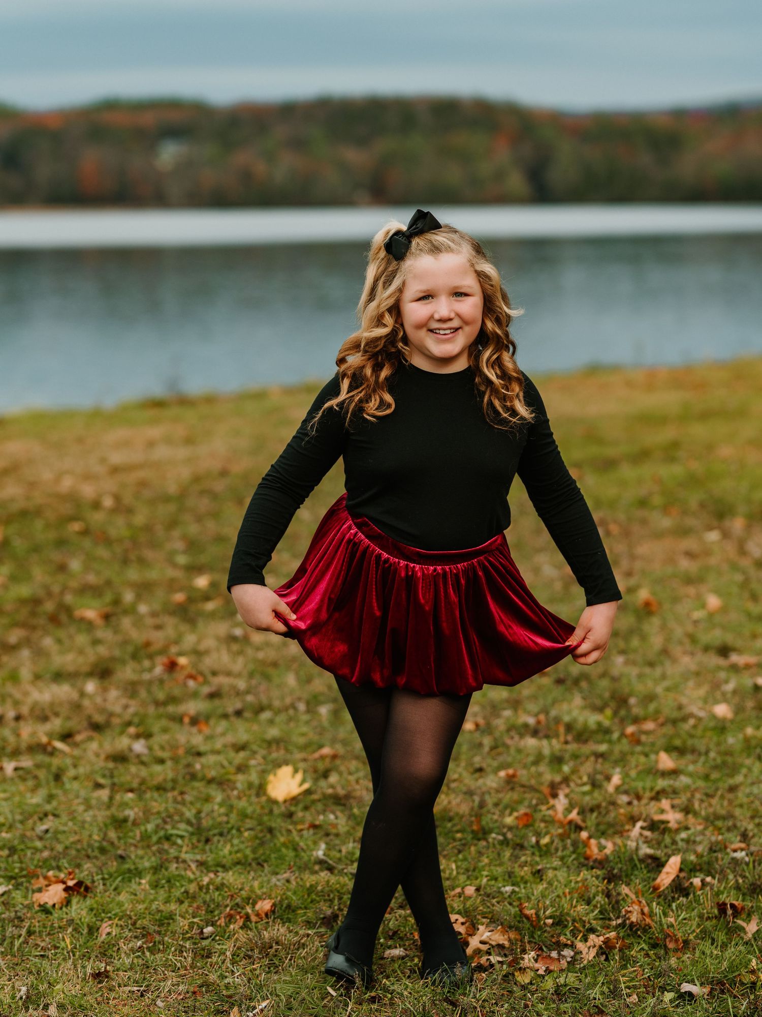 A child in a red velvet dress plays joyfully in fallen autumn leaves by a lakeside on a crisp fall day.
