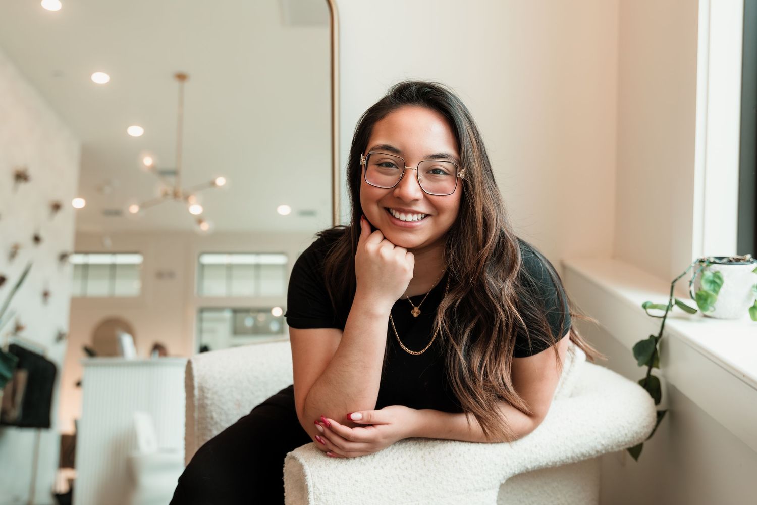 Young woman with glasses and long hair smiling warmly while seated in a bright modern interior space.