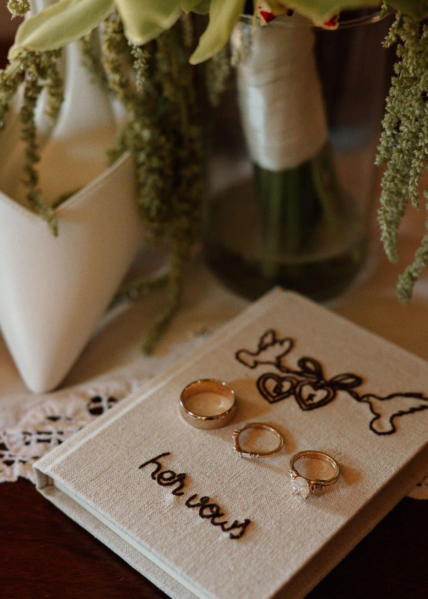 Gold wedding rings resting on a lace-covered vow book beside bridal bouquets with greenery.