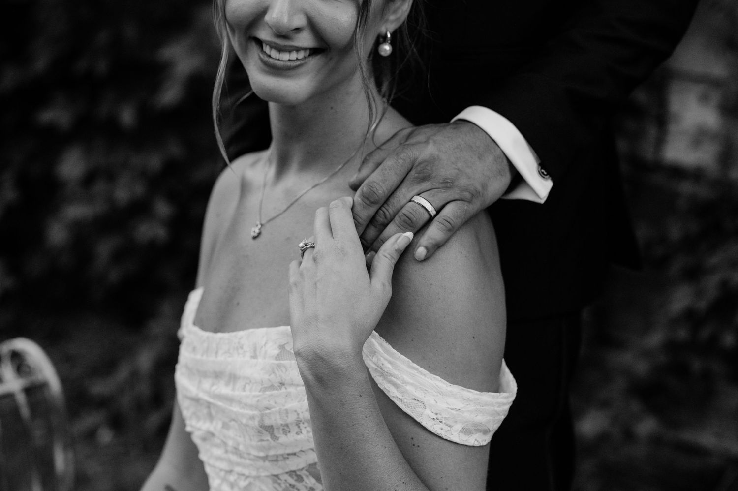 Black and white wedding photo of bride in off-shoulder dress with groom's hand on her shoulder.