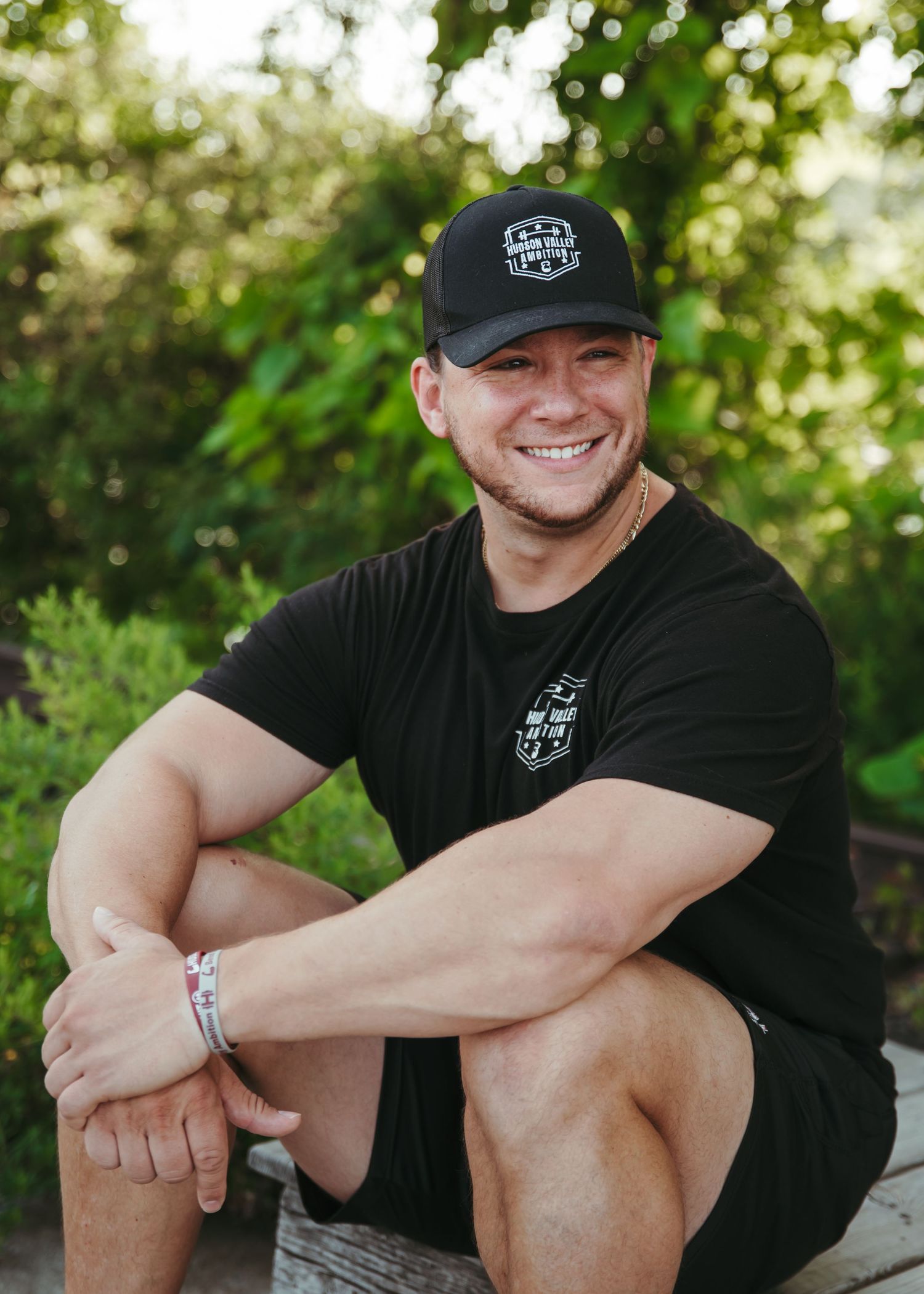 A person wearing a black t-shirt and baseball cap sits casually outdoors with greenery in the background.