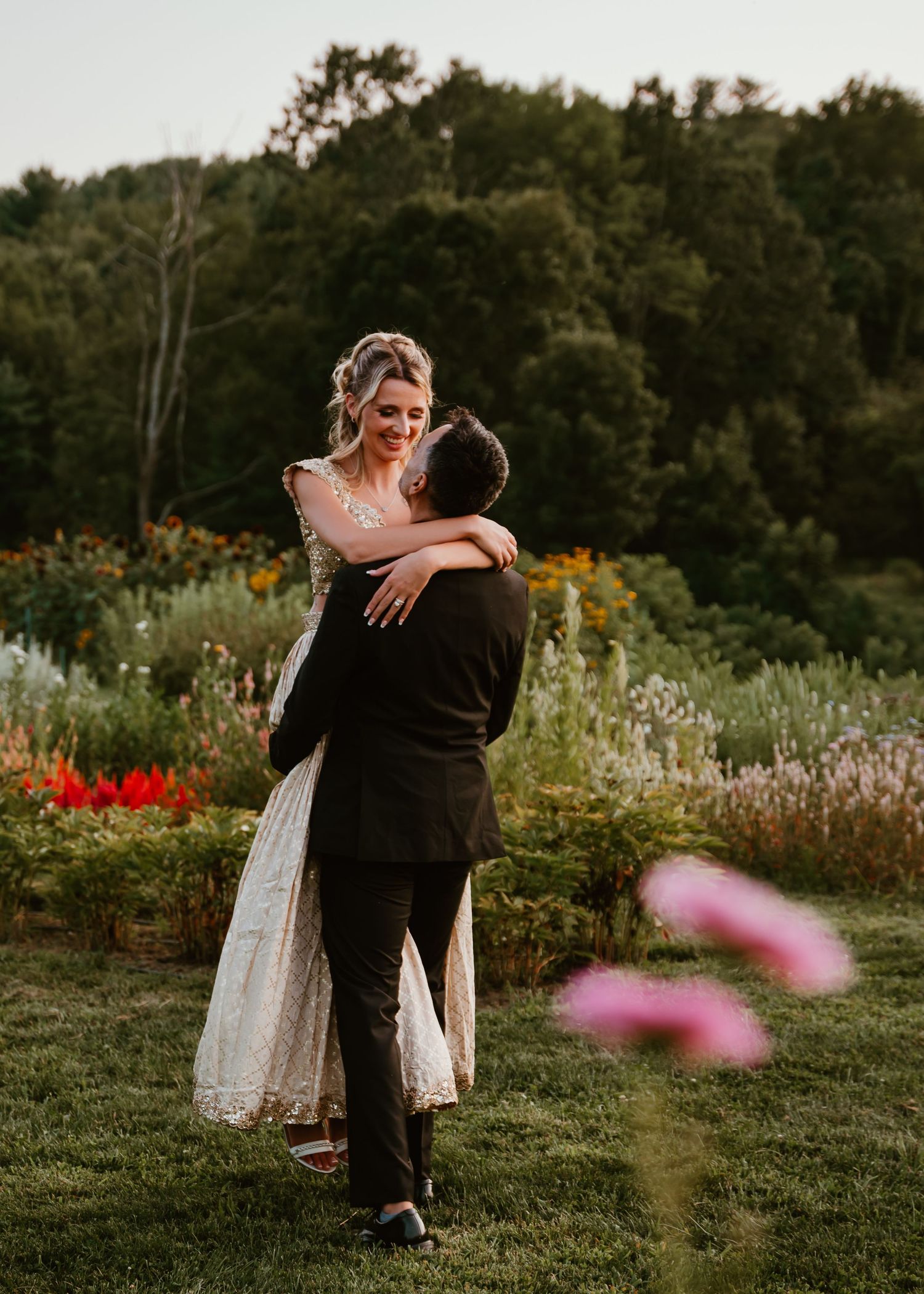 A couple embraces in a garden meadow at sunset with pink wildflowers in the foreground and trees in the background.