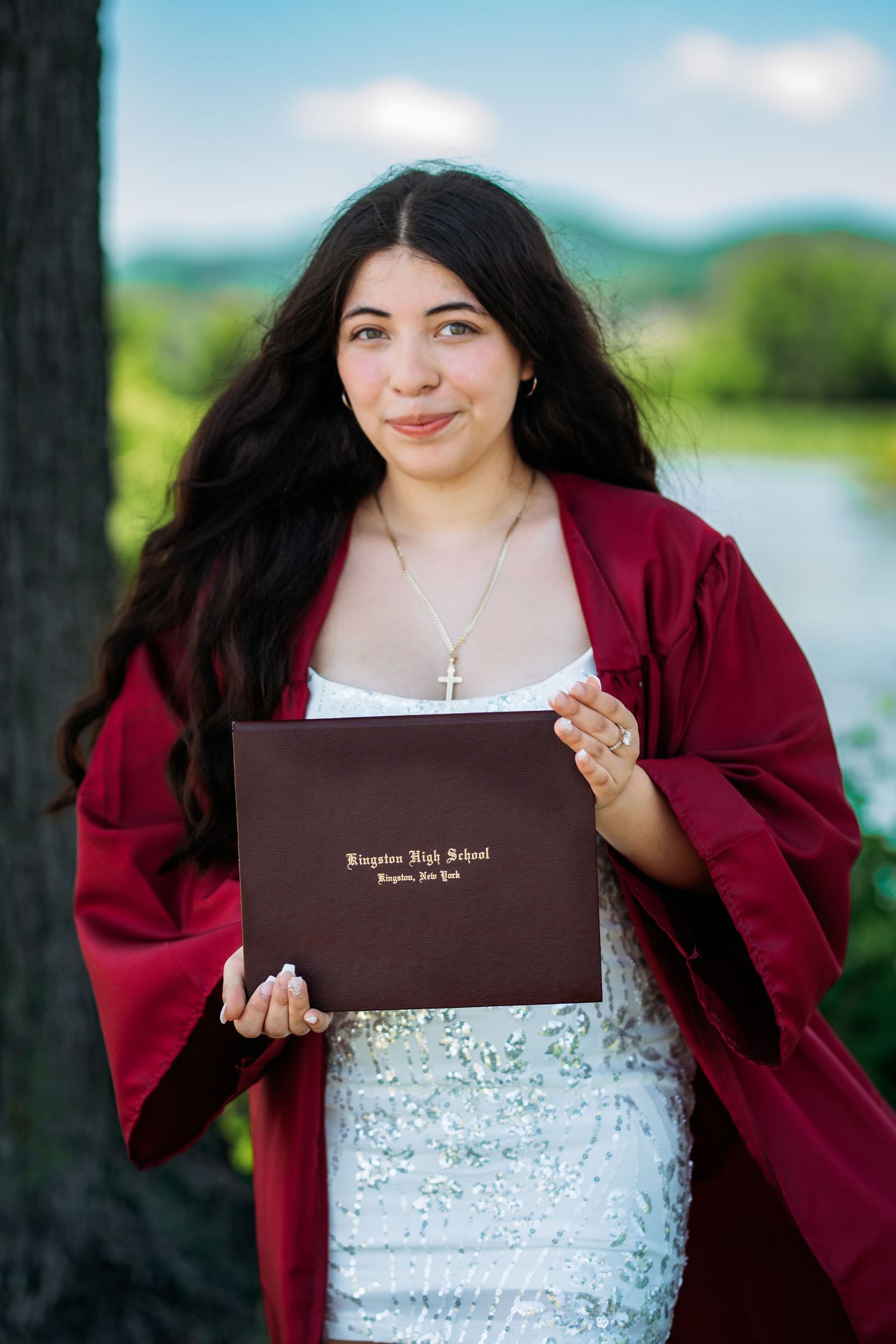 Young woman in red graduation gown holds Kingston High School diploma outdoors near water.