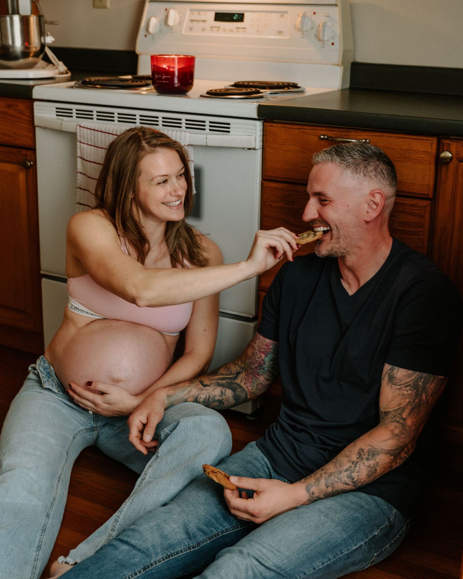 Two people sit casually on a kitchen floor in front of appliances while sharing a relaxed moment together.