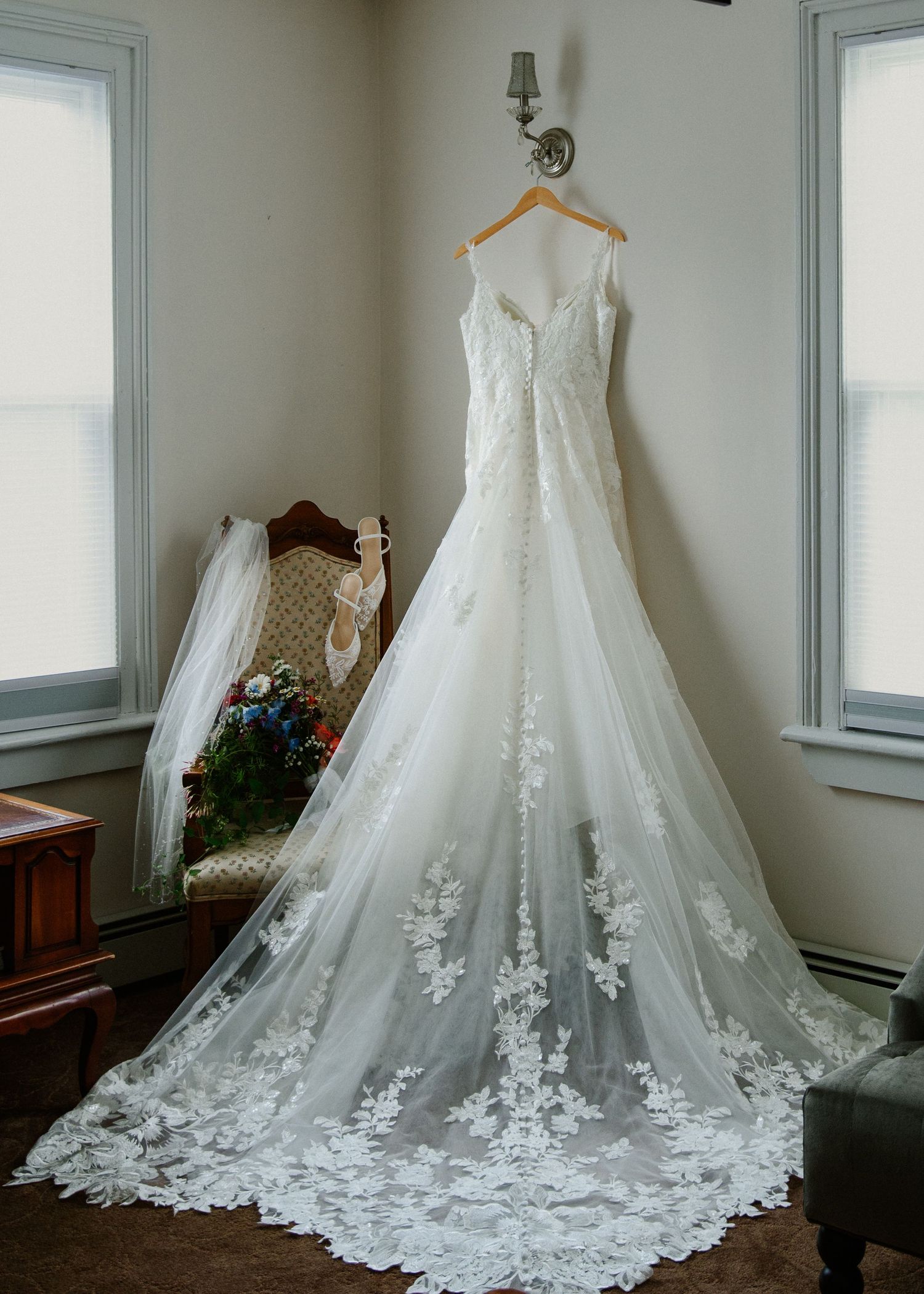 Elegant white lace A-line wedding dress with floral appliques and long train hanging on a wooden hanger in a bright room.
