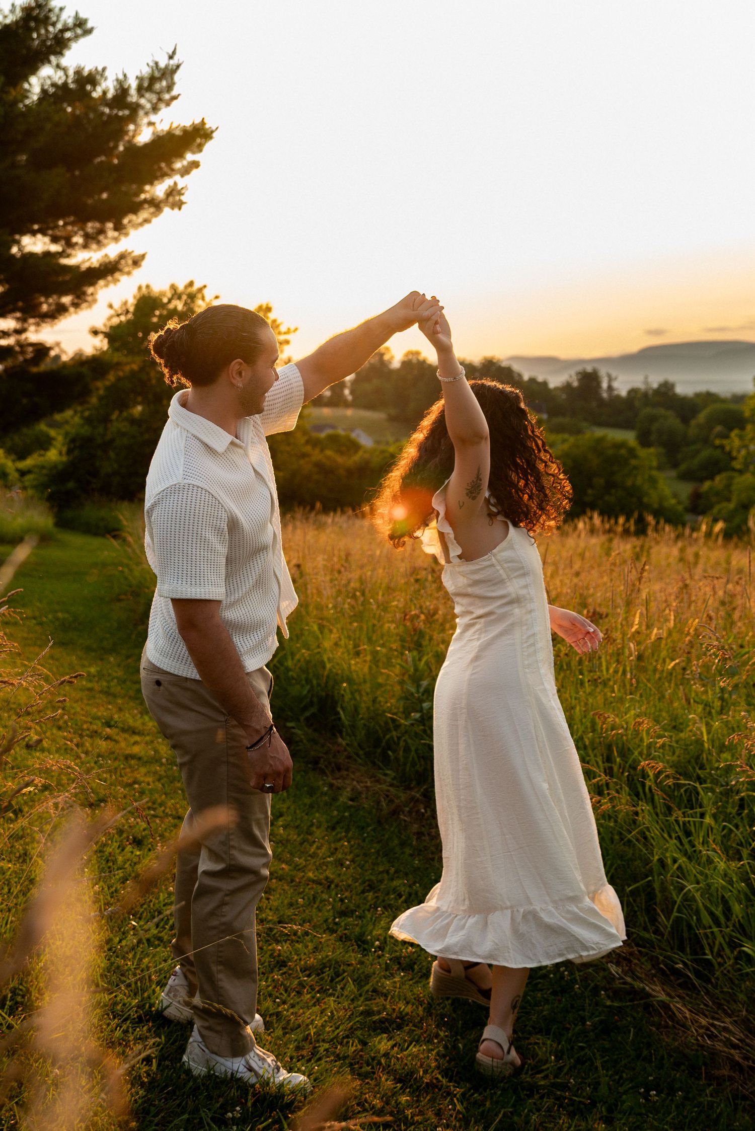 Couple dancing at sunset in a golden field, man spinning woman in white dress, romantic outdoor engagement photo.