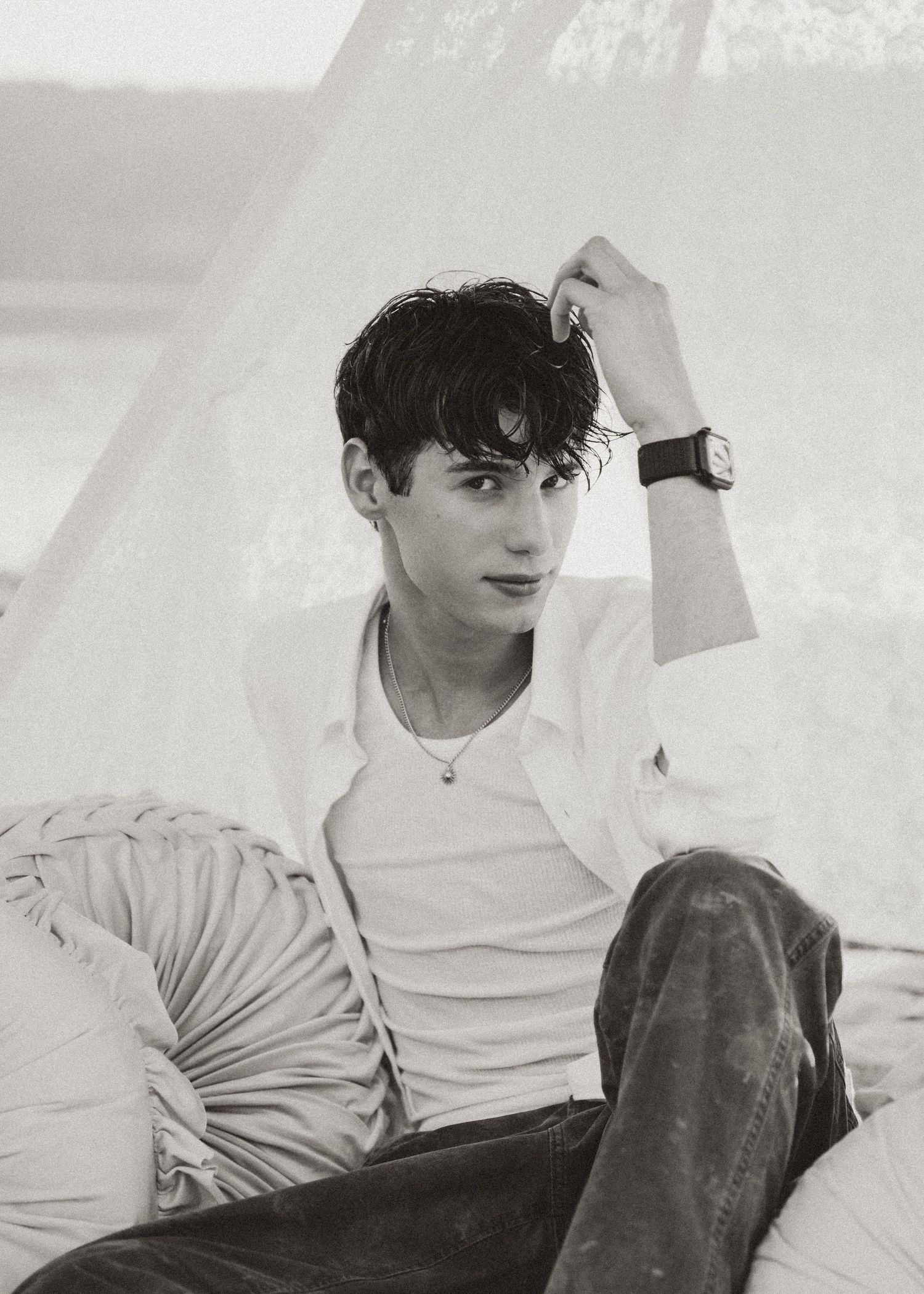 Young man in white shirt and jeans sits relaxed, hand in dark hair, wearing a watch, in a dreamy black and white photo.