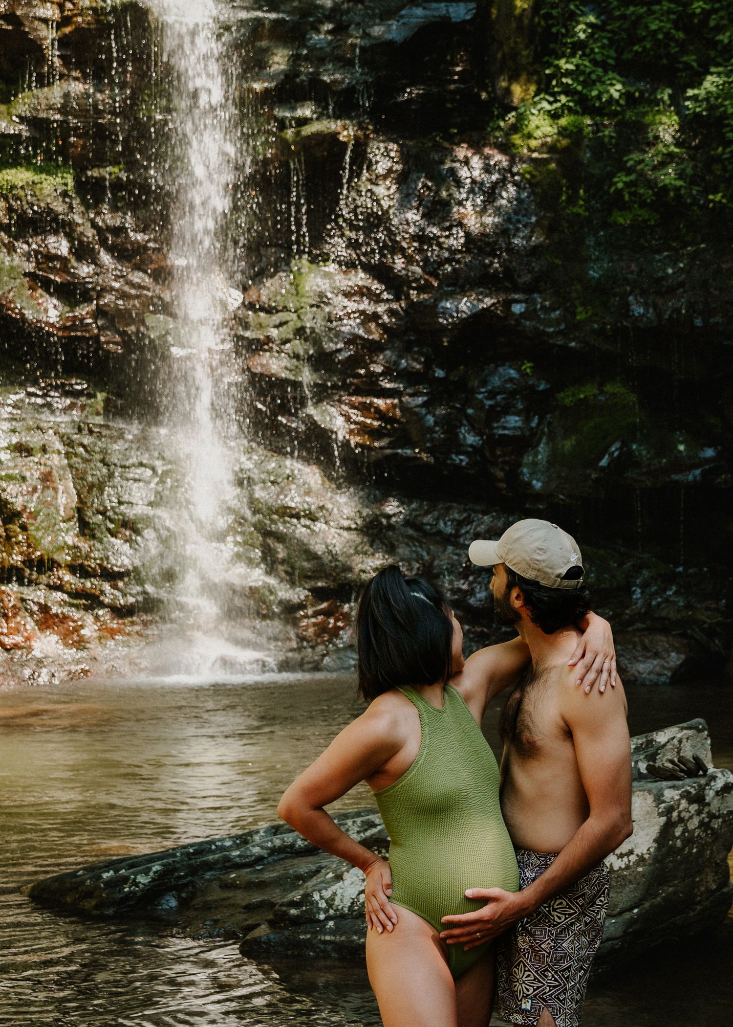 Couple embracing near a waterfall, woman in green swimsuit with baby bump, man in shorts, lush forest backdrop.