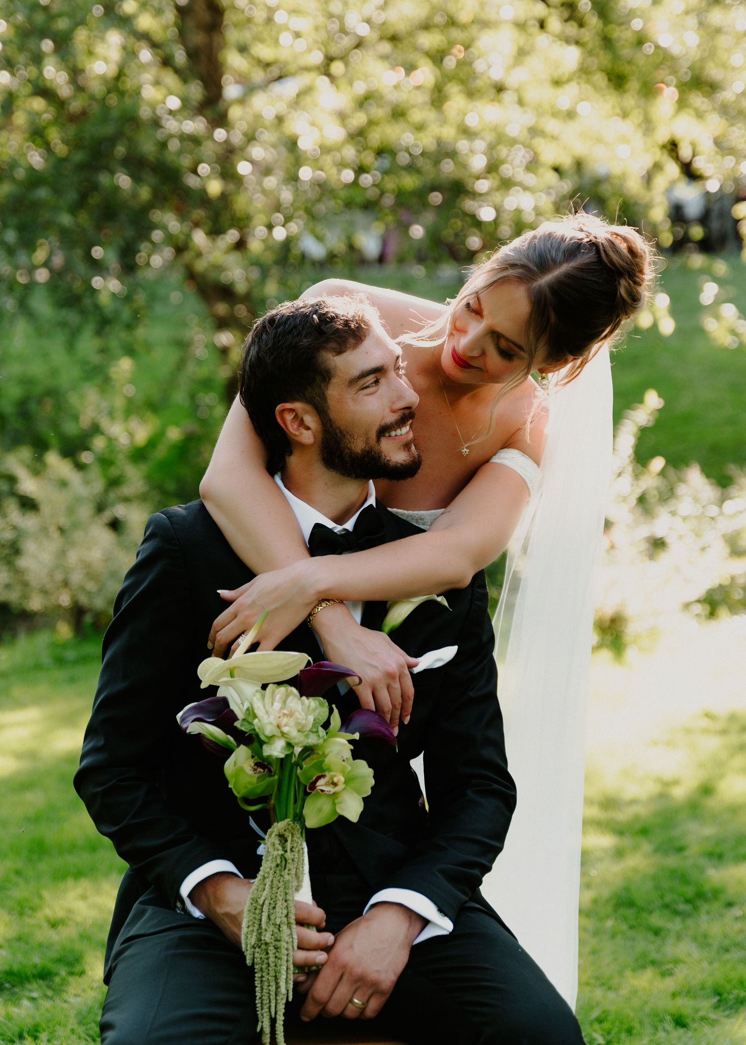 A groom in a black tuxedo kneels outdoors as his bride in a white gown embraces him, holding a green bouquet.
