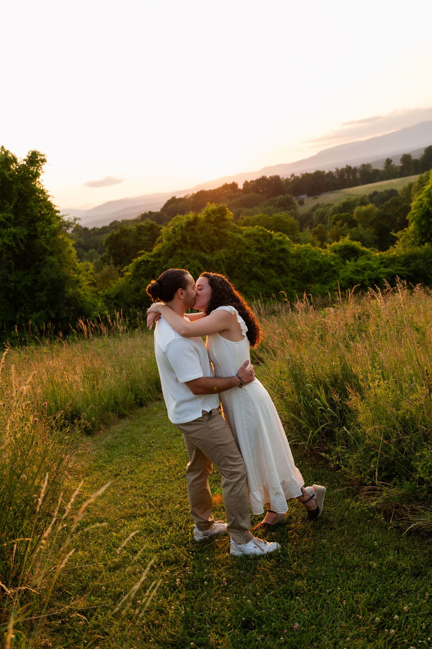 Couple sharing a romantic kiss at golden hour in a lush green meadow with mountain views.