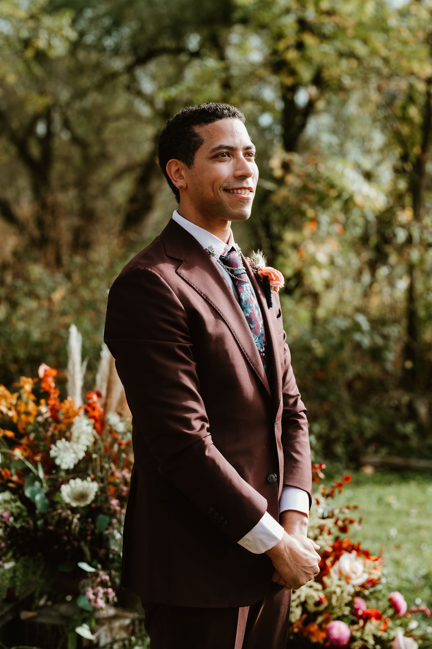 A groom in a burgundy suit smiles warmly while standing in a garden setting with autumn flowers.