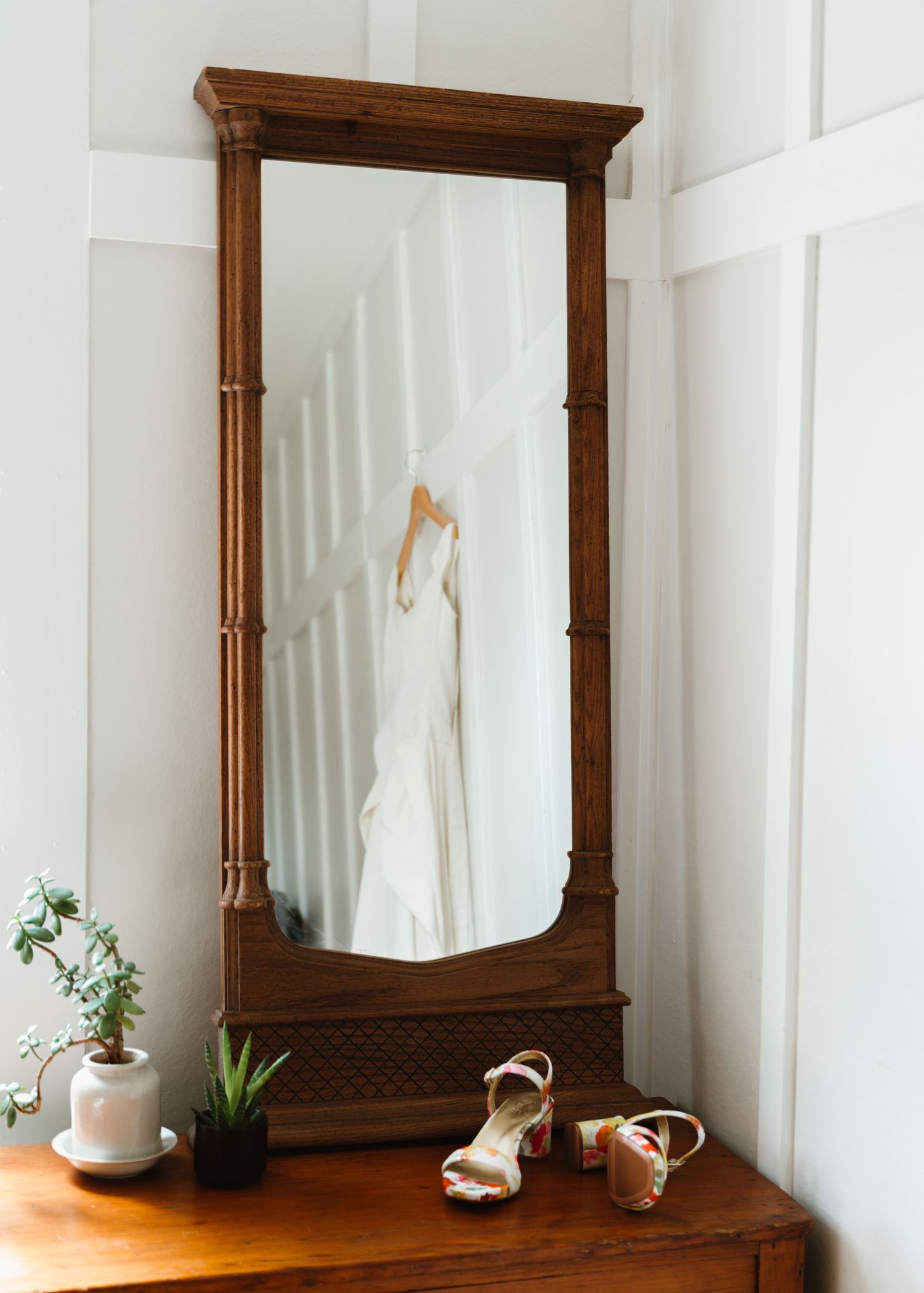 A vintage wooden dresser with mirror displays decorative items and plants against a white wall in a minimalist setting.