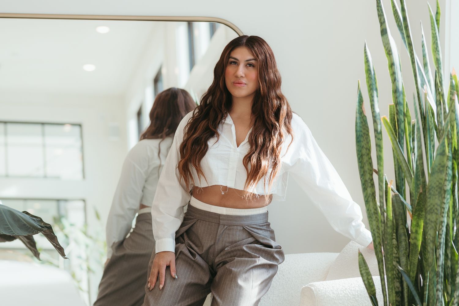 Woman with long brown hair wearing white crop top and gray trousers posing near mirror and indoor plant.