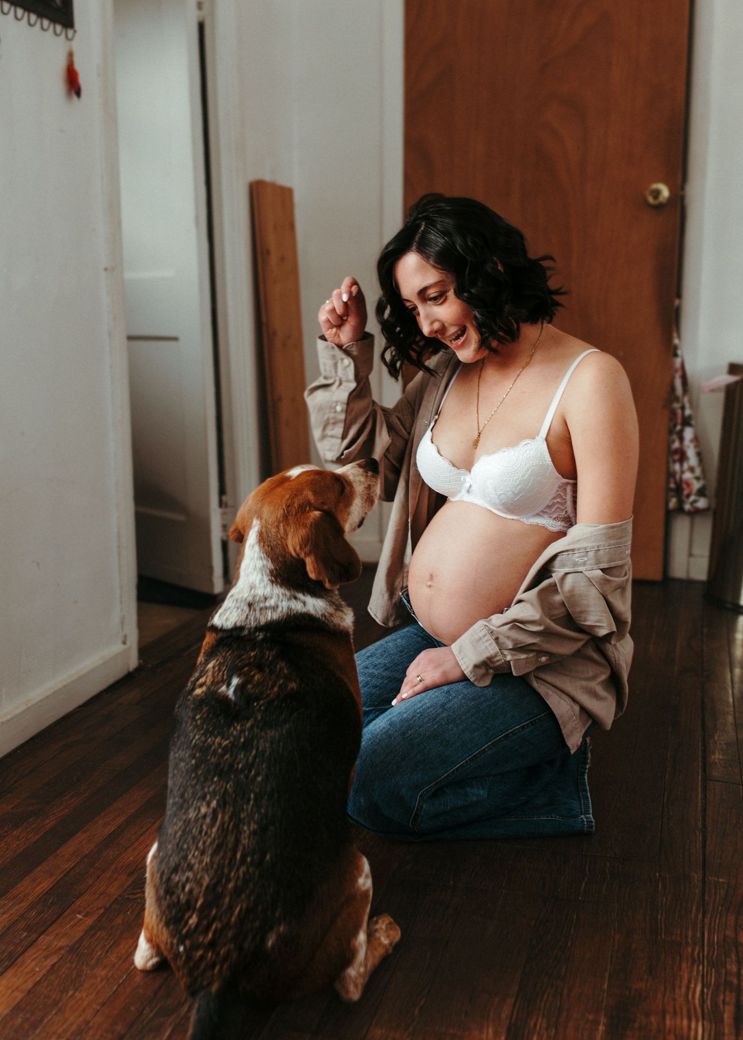 Pregnant woman in white bra and jeans sits on wood floor smiling at her beagle dog indoors.