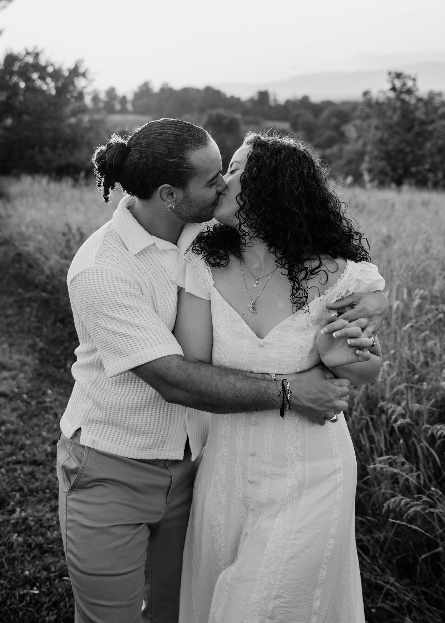 Black and white photo of a couple kissing in a field, woman in white dress, man in striped shirt.