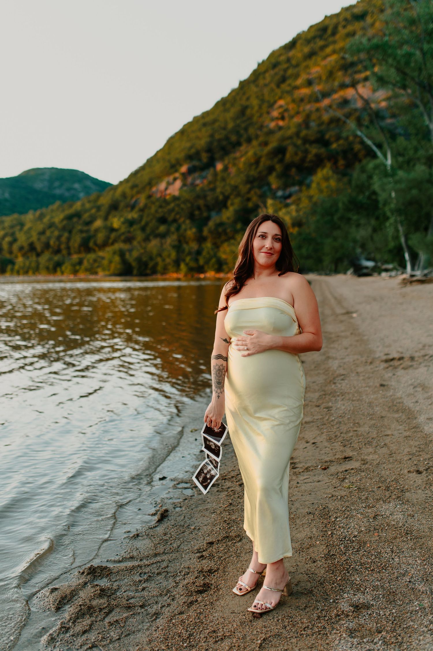 Pregnant woman in sage green tube dress holding ultrasound photos on a lakeside beach with green mountains behind her.