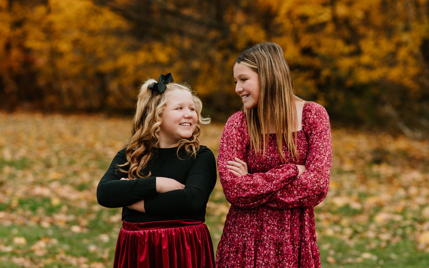 Two people in red and black dresses share joyful moments among golden autumn leaves in an outdoor setting.