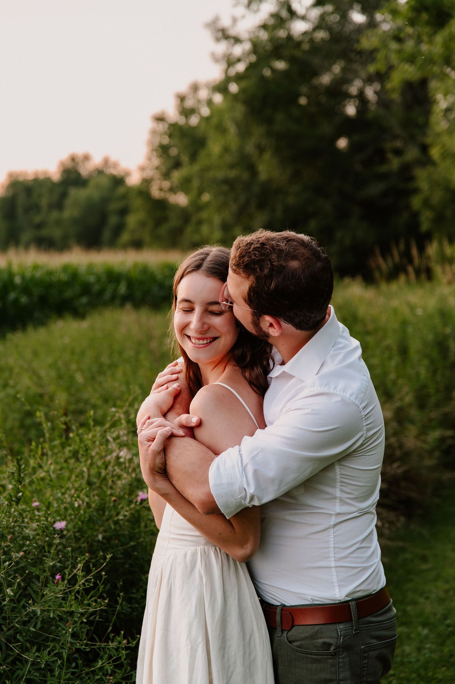 A couple embraces in a lush green field at golden hour, the man kissing her cheek as she smiles joyfully.