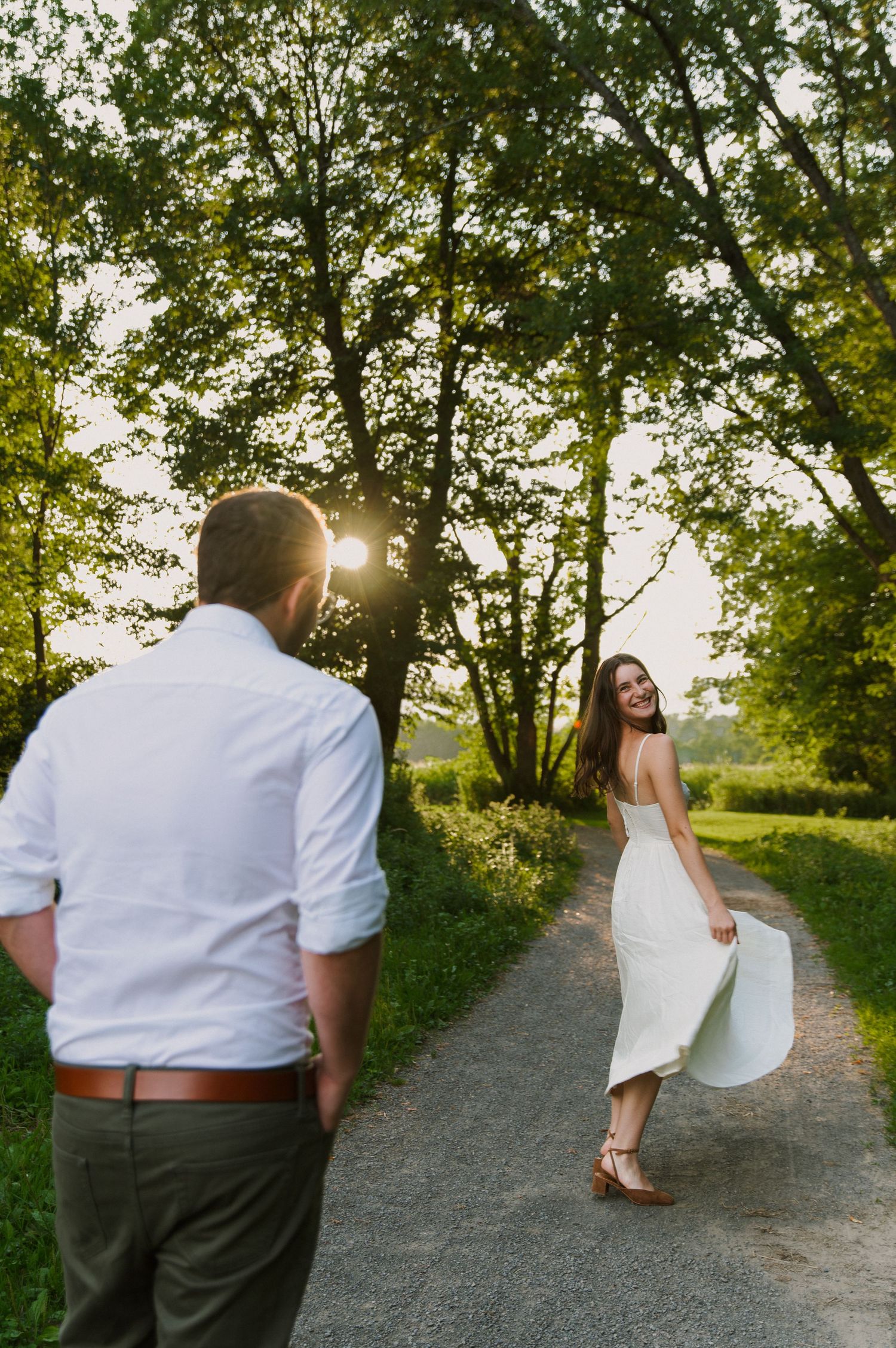 Couple on a sunlit gravel path, woman in white dress smiling back at man in white shirt among lush green trees.