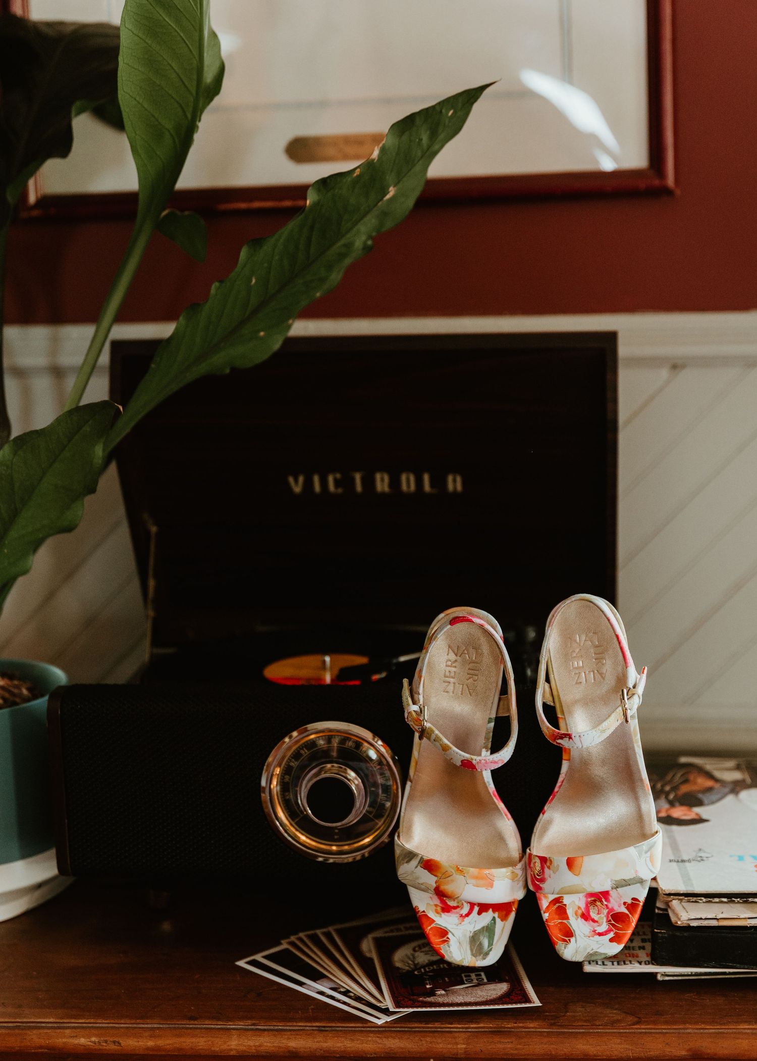 Floral patterned wedding shoes displayed next to a Victrola record player and green plant on dark wooden surface.