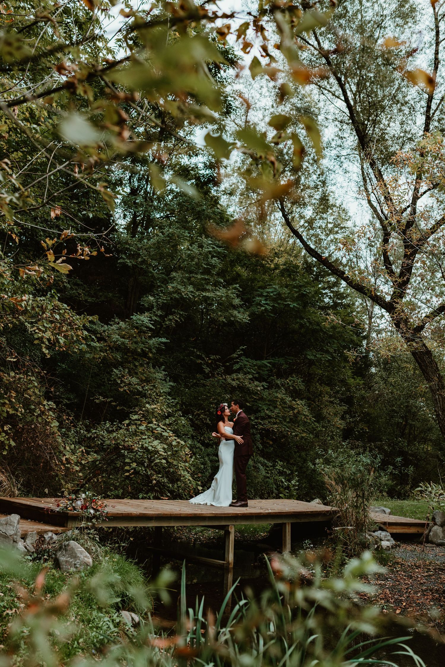 Couple sitting together on rustic wooden bench surrounded by lush green trees and foliage in romantic garden setting.