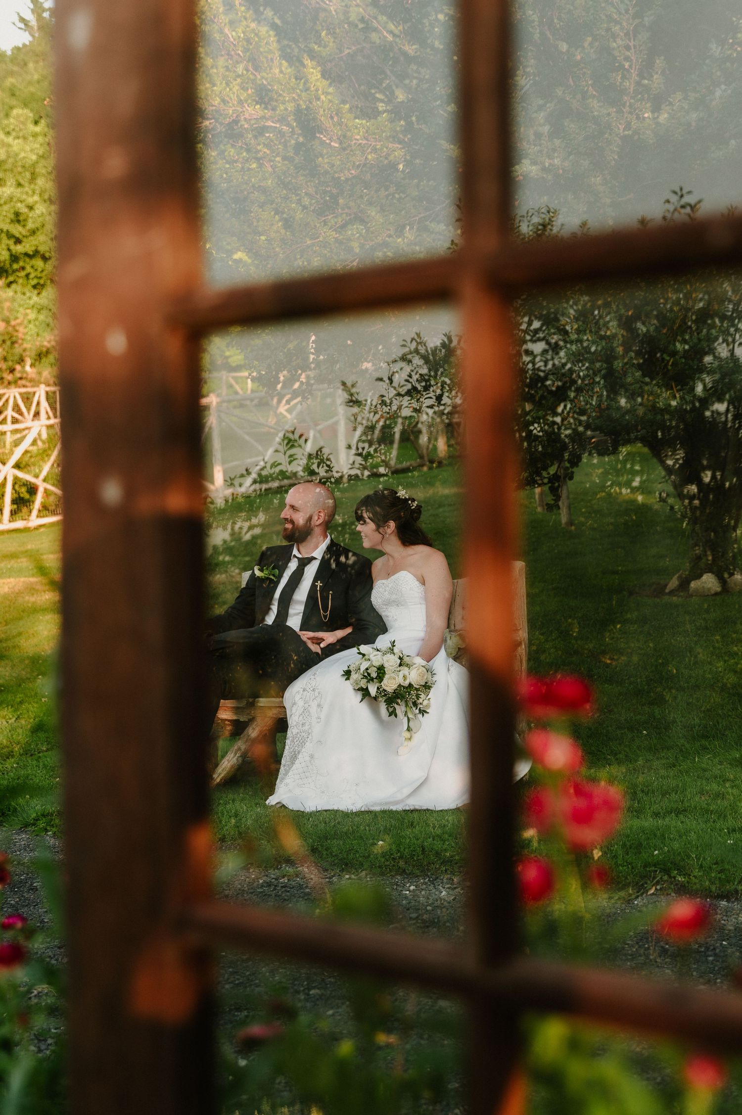 Bride and groom sitting together in lush garden, viewed through rustic window frame, bride holding white bouquet.