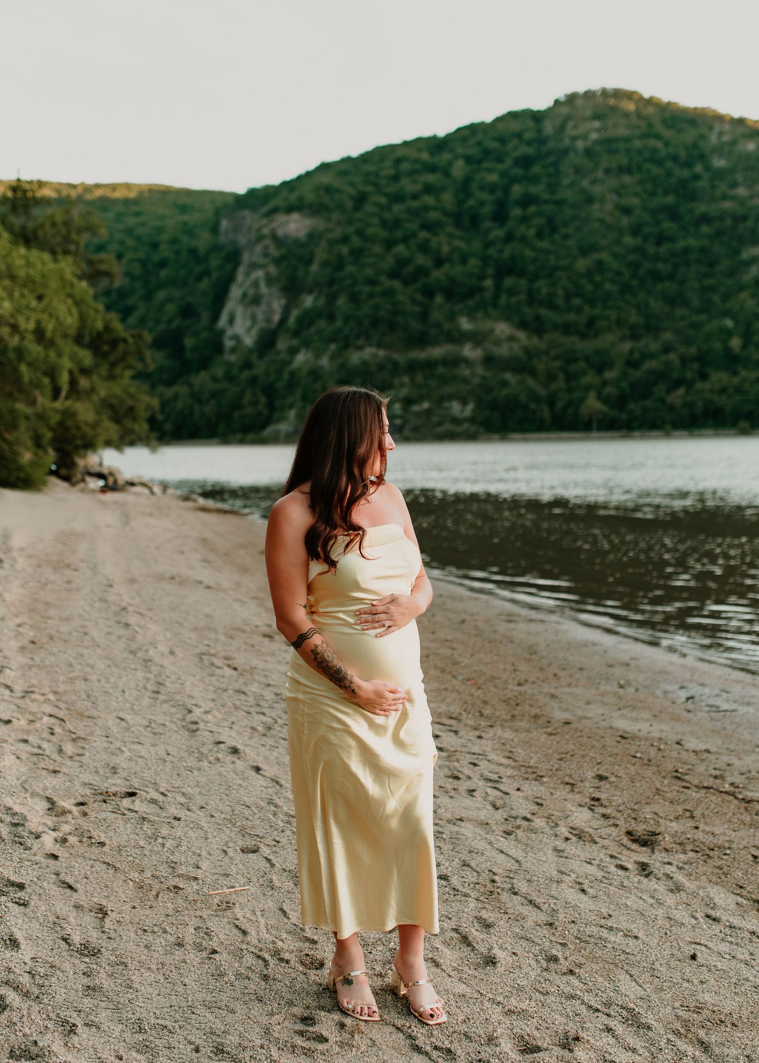 Pregnant woman in yellow satin dress cradling baby bump on sandy beach with mountain backdrop.
