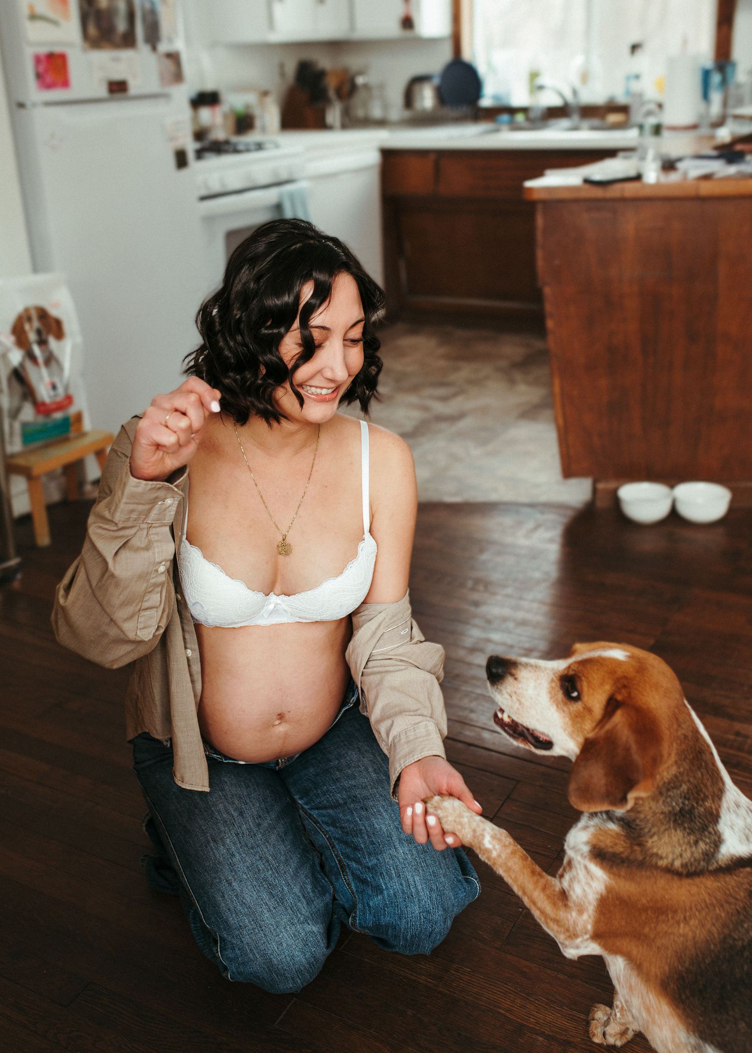 Pregnant woman in white bra and jeans kneeling on wood floor, smiling while beagle dog shakes her hand in kitchen.