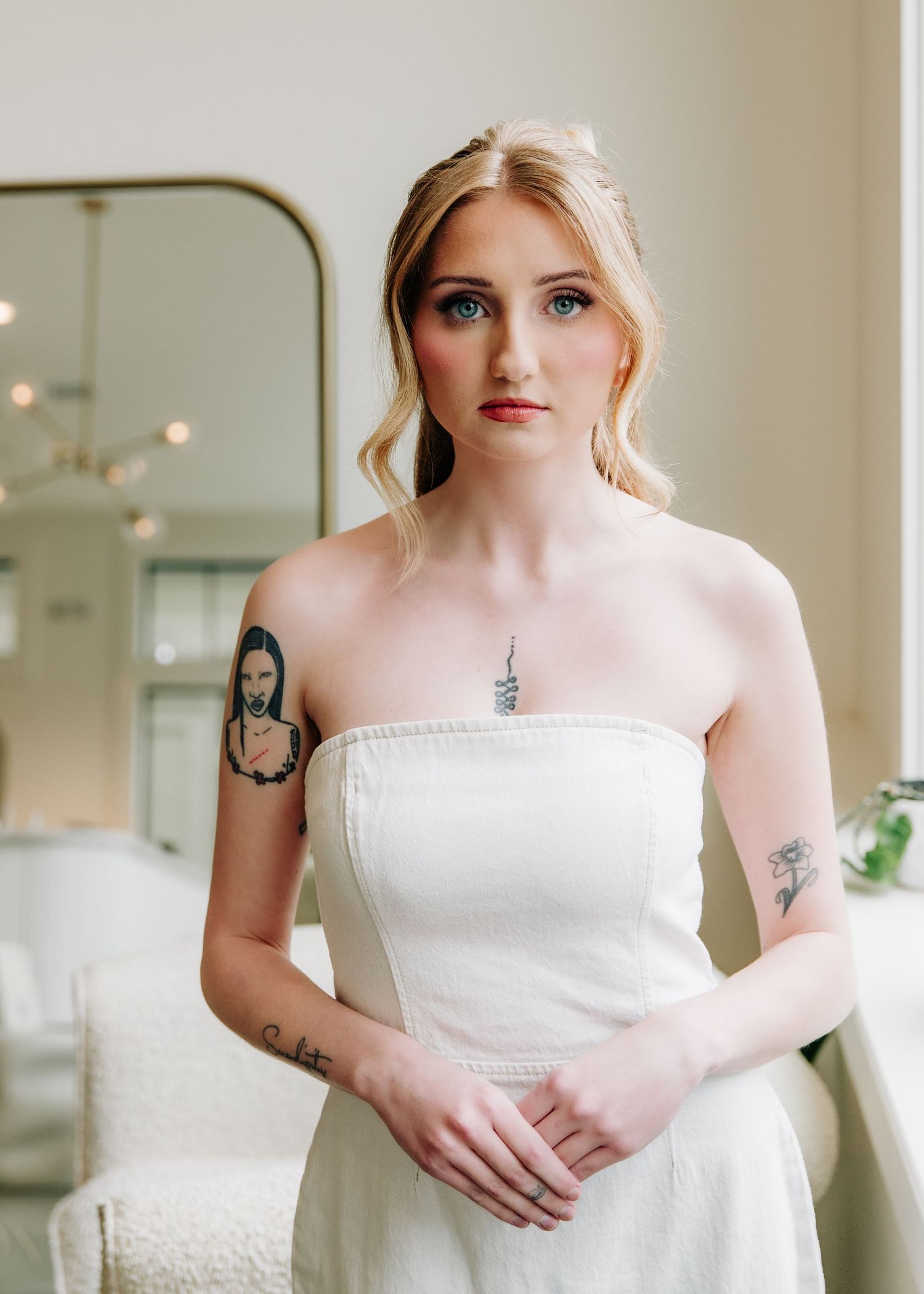 Blonde bride in strapless white dress with tattoos poses elegantly in a softly lit room with a gold mirror.