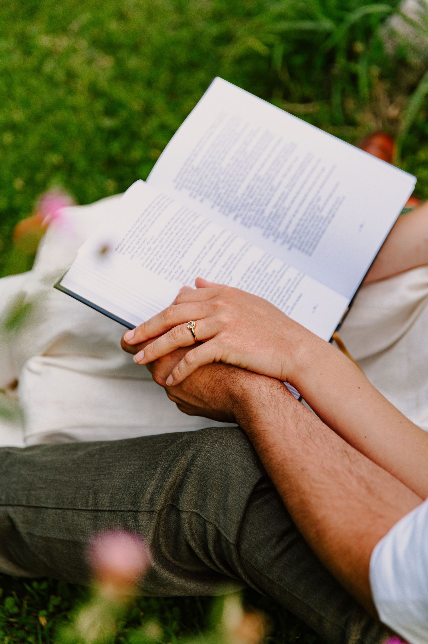 Couple sitting on grass reading a book together, hands intertwined with a gold ring visible.
