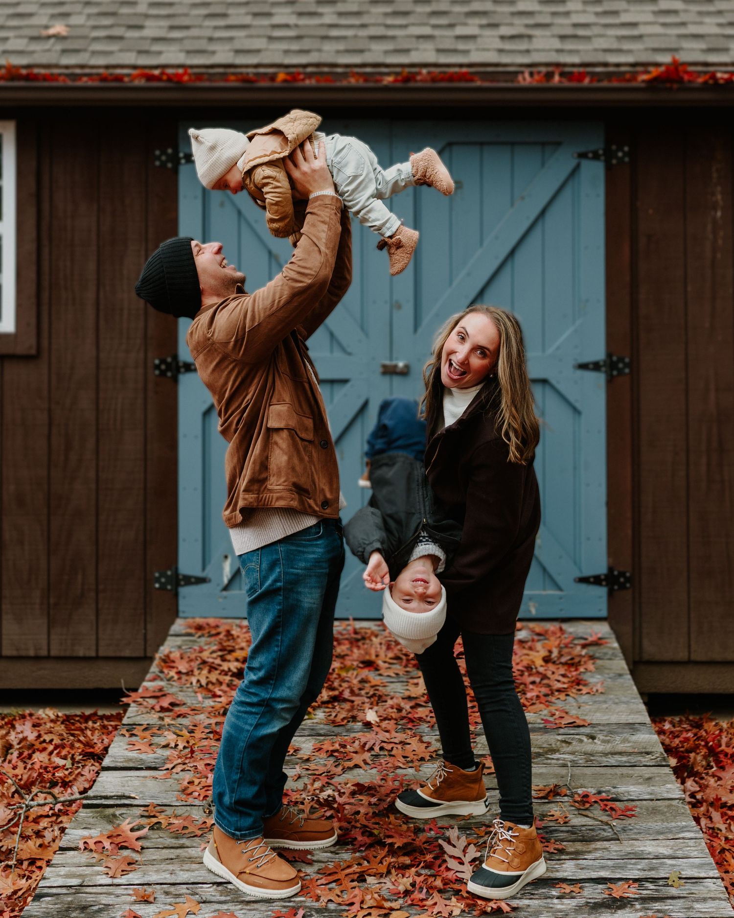 A family enjoying an autumn day together in front of a rustic barn door with fallen leaves scattered on the ground.