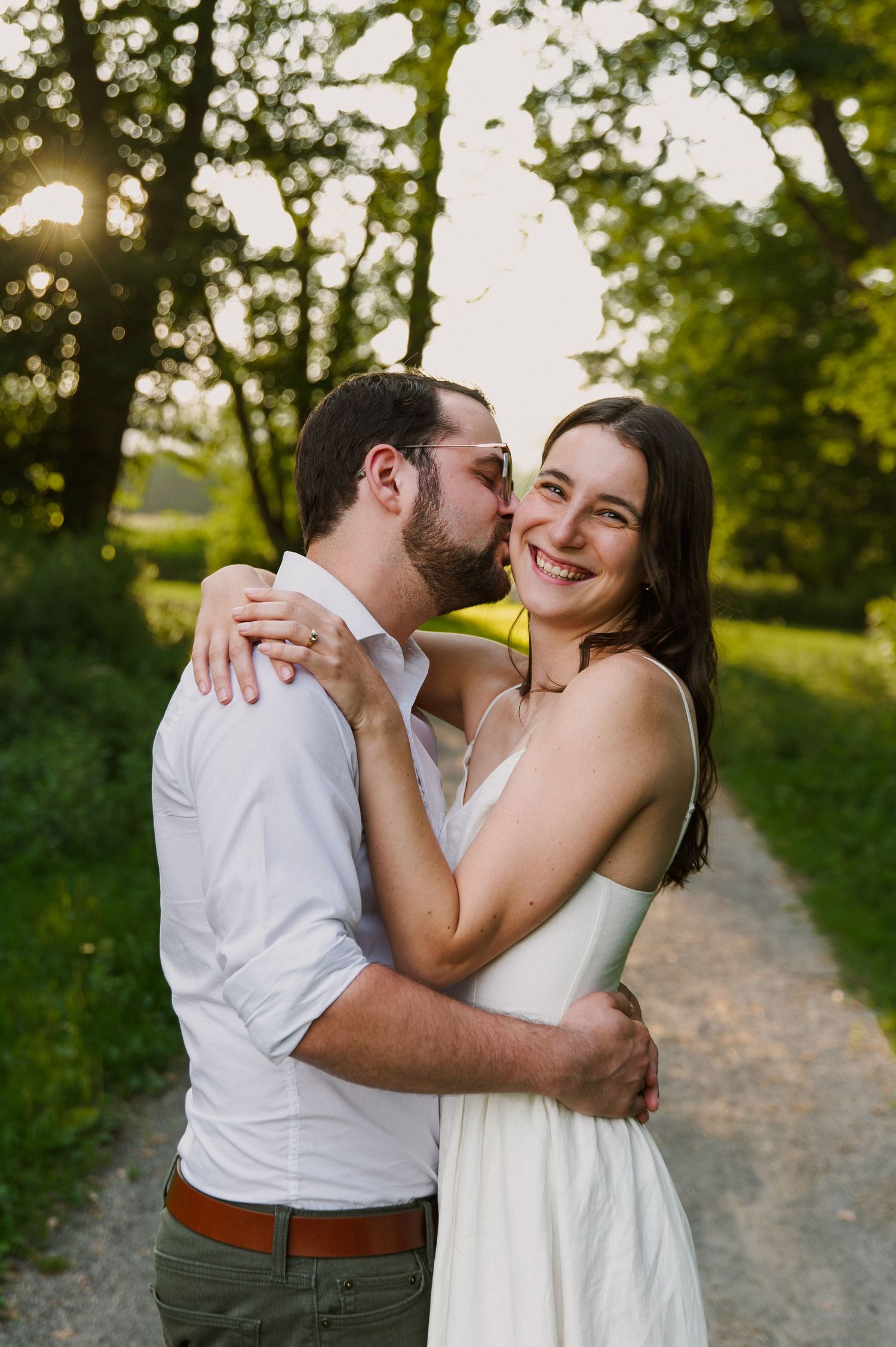 A couple shares a tender kiss at golden hour on a tree-lined path, the woman smiling in a white dress.
