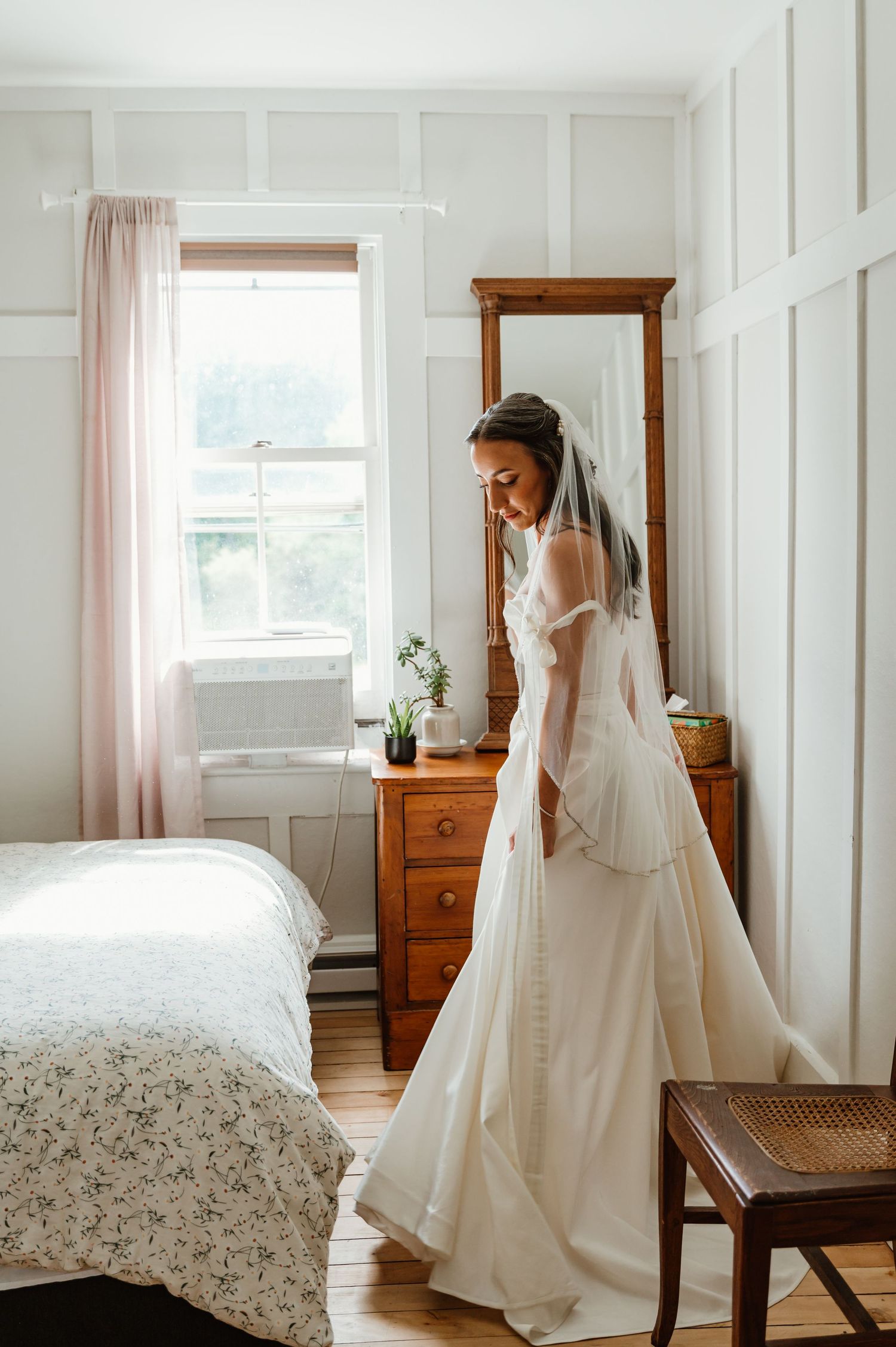 A bride in an elegant white wedding gown stands in a sunlit bedroom with vintage wooden furniture and floral bedding.